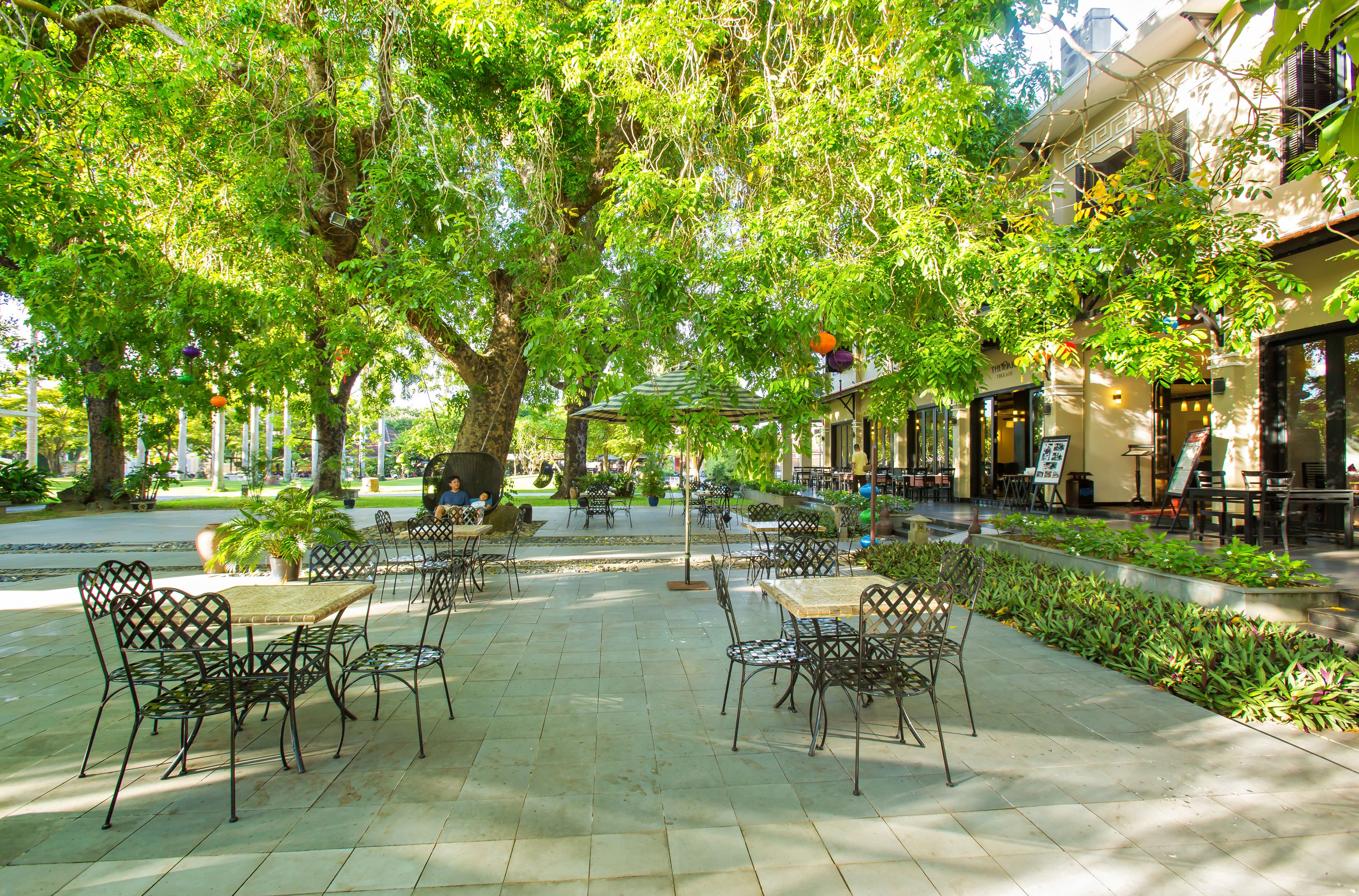An outdoor café with tables and chairs, surrounded by trees and greenery.
