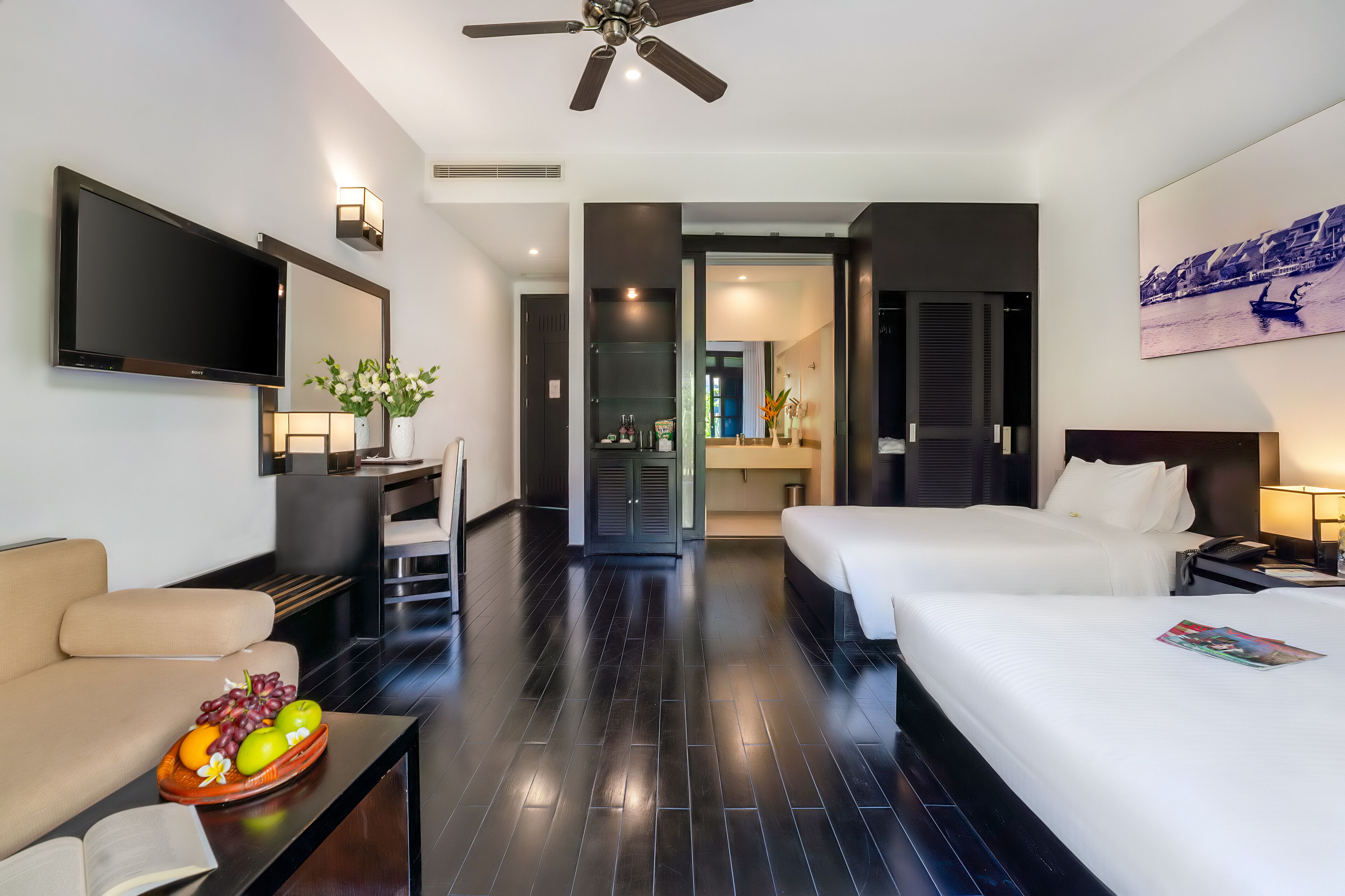 A hotel room with two beds, a TV, a desk, and a seating area with a fruit bowl.