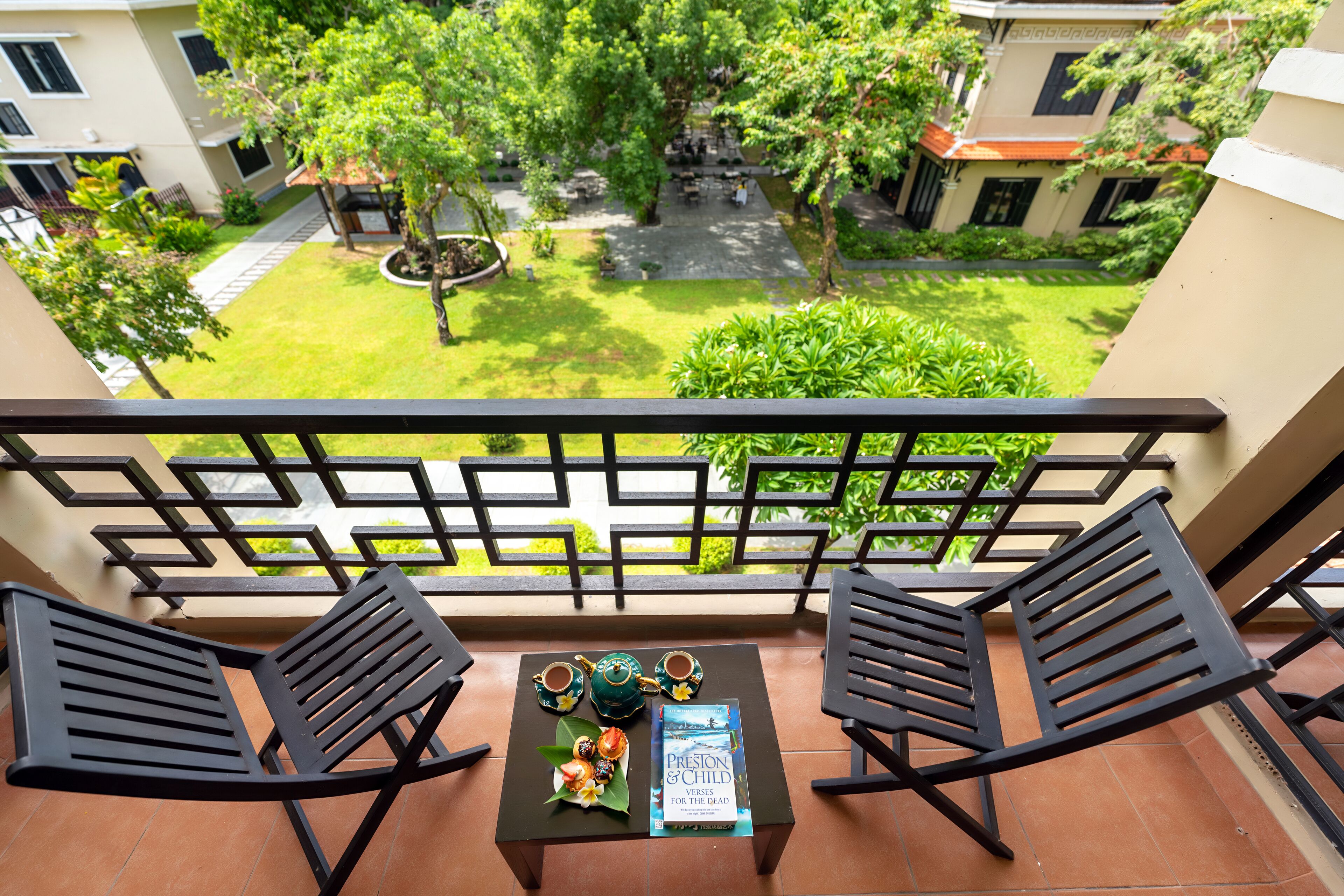 A balcony with two black chairs, a small table with a book and teacups, and a view of a green courtyard with buildings in the background.