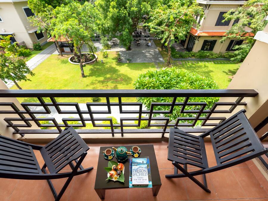 A balcony with two black chairs, a small table with a book and teacups, and a view of a green courtyard with buildings in the background.