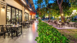Un área de comedor al aire libre con mesas y sillas, un sendero bordeado de vegetación y un edificio con amplias ventanas e iluminación exterior.