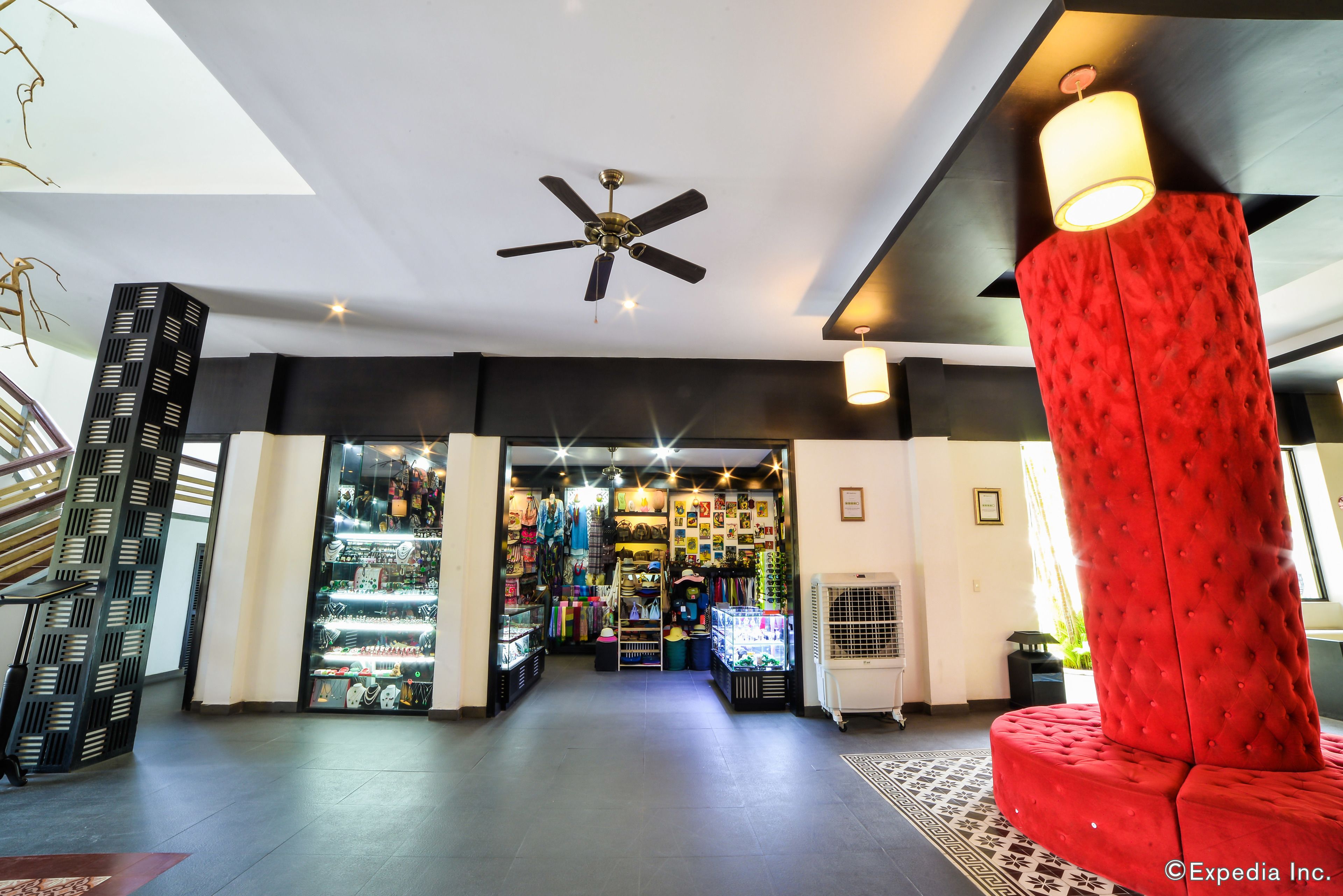 Un vestíbulo de hotel con ventilador de techo, una zona de estar tapizada en rojo y una tienda bien iluminada con diversos artículos a la vista.