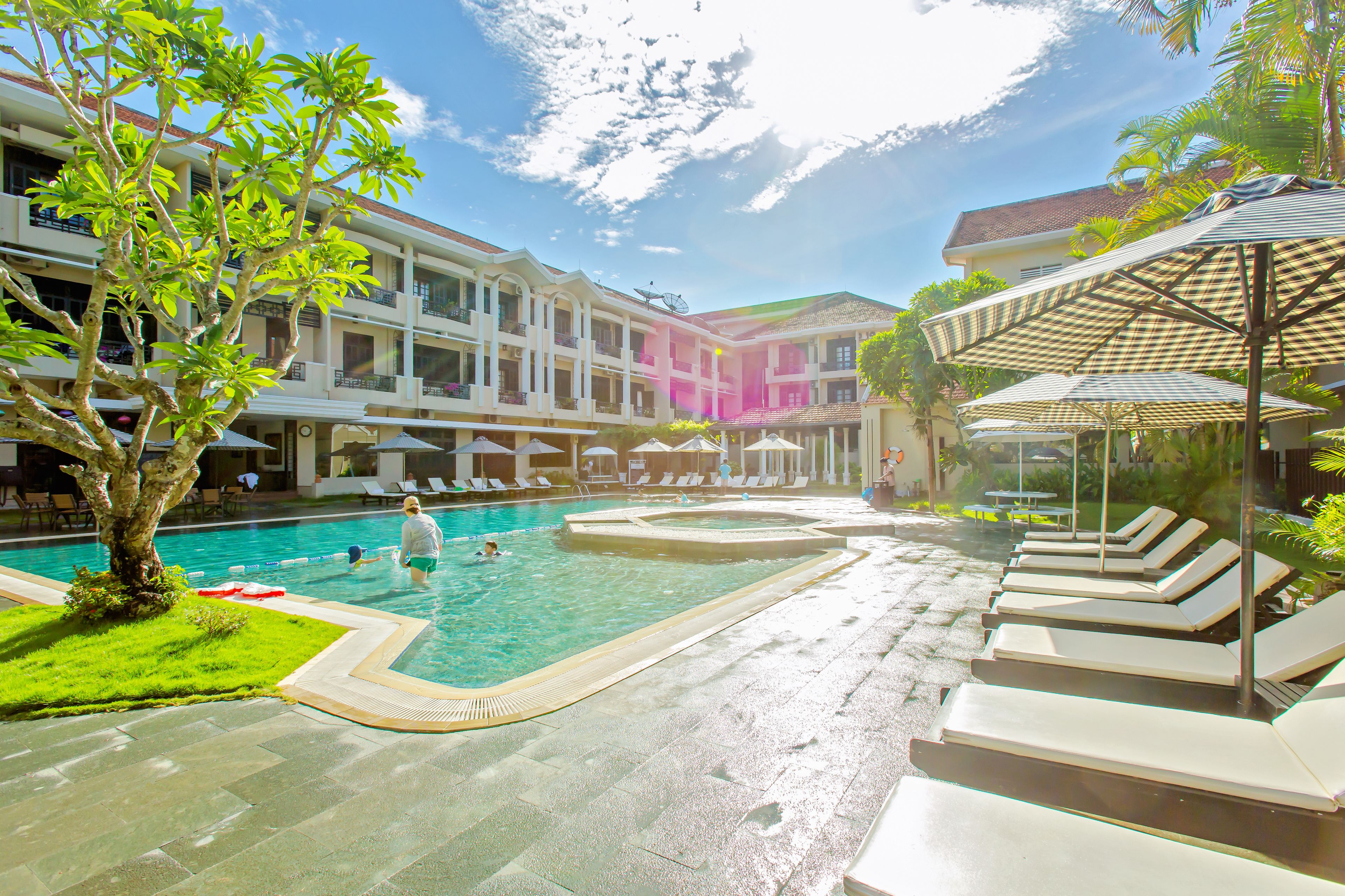 Ein Hotelpoolbereich mit mehreren Liegestühlen und Sonnenschirmen, umgeben von einem Gebäude mit Balkonen.