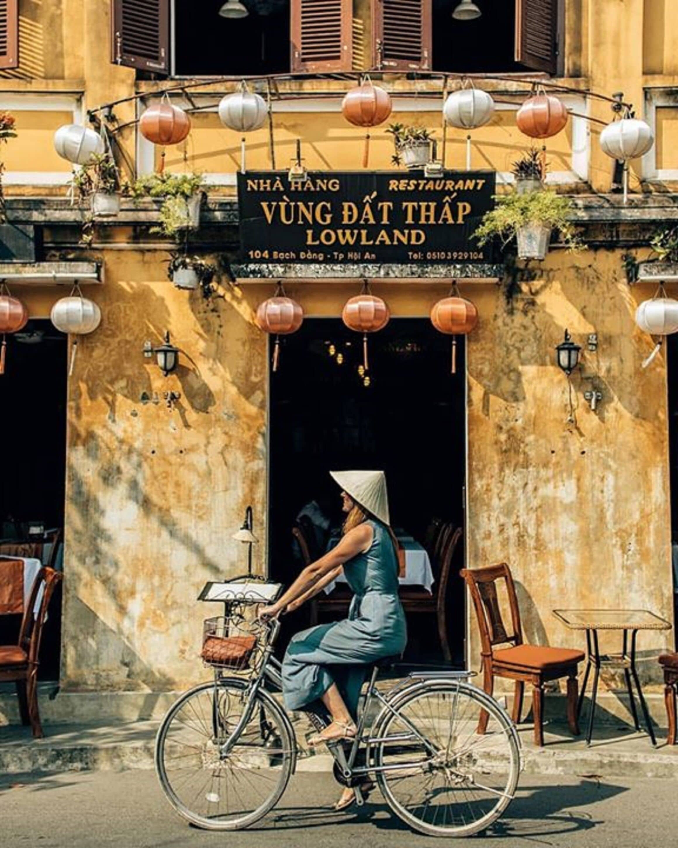 En kvinna cyklar förbi en restaurang som heter ”NHA HANG VUNG DAT THAP LOWLAND” och som har uteservering och lyktor.