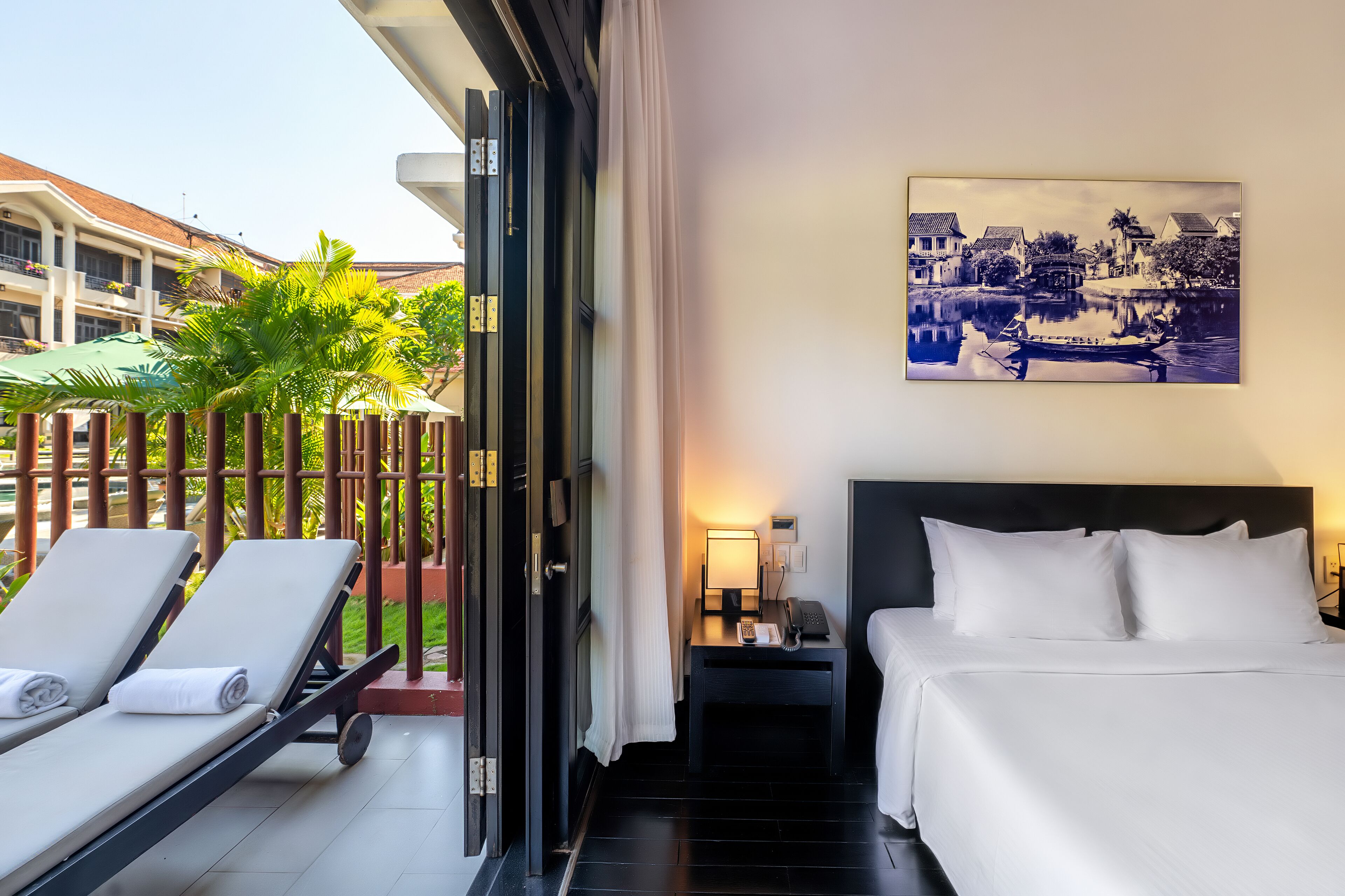 A hotel room with a bed, a nightstand, a lamp, and a view of a courtyard with lounge chairs and a pool.
