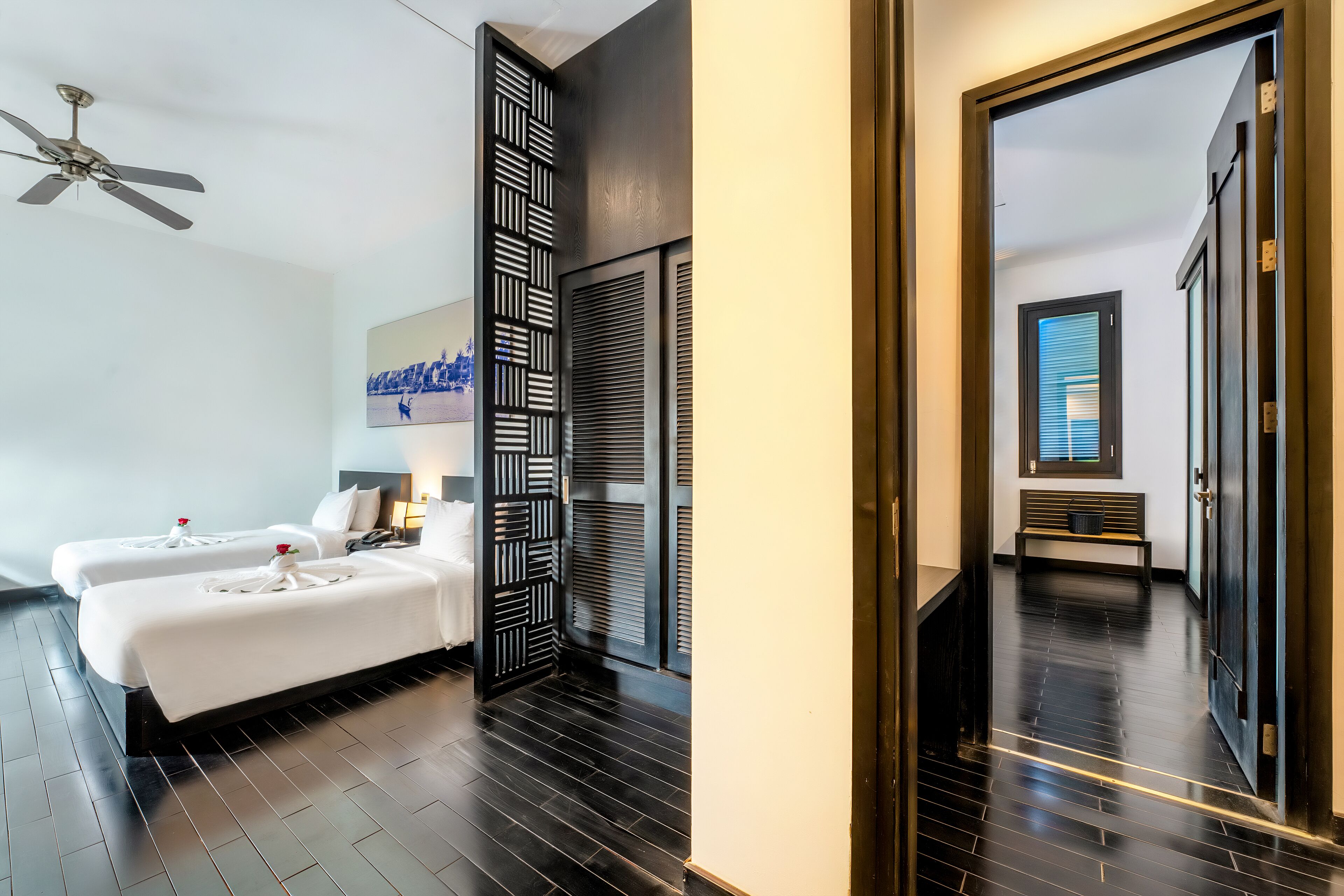 A modern hotel room with two beds, a ceiling fan, a decorative wall panel, and a bench in the hallway.