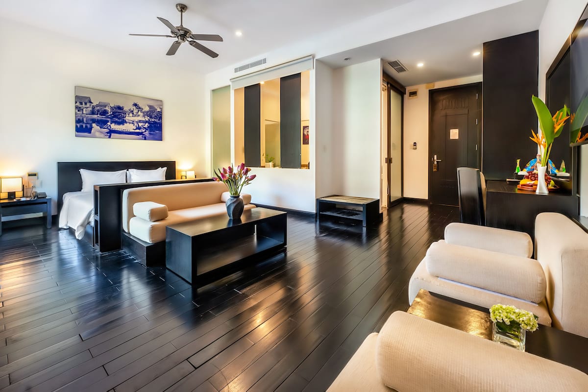 A modern hotel room with a dark wooden floor, a sofa, a coffee table, a bed with white bedding, and a large mirror.