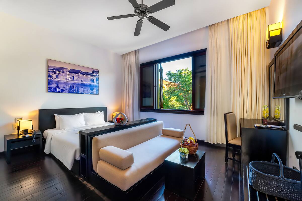 A modern hotel room with a large bed, a sofa, a desk with a lamp, a TV, and a window with curtains.