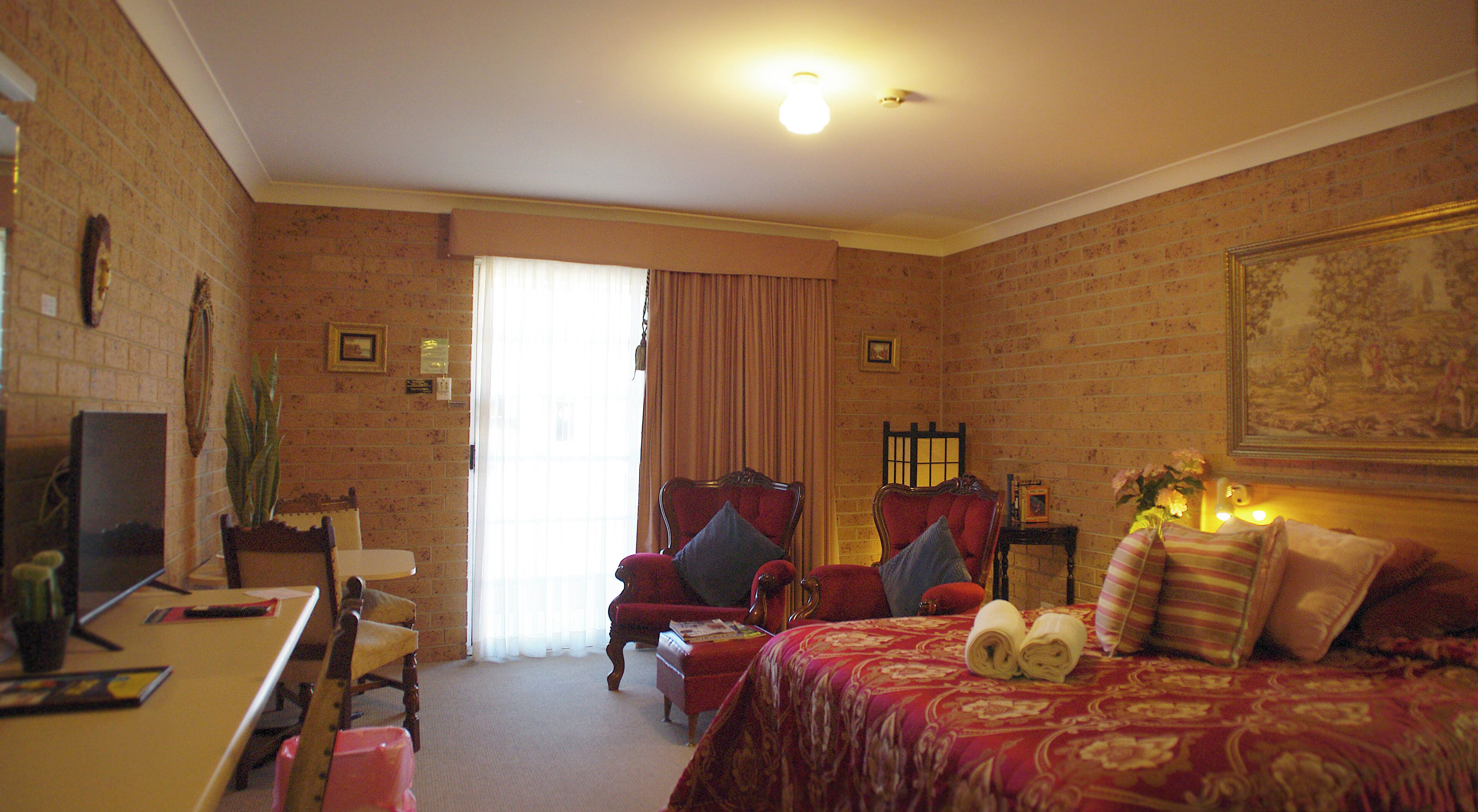 A hotel room with a brick wall, a large bed with red bedding, two armchairs, a small table, and a television.