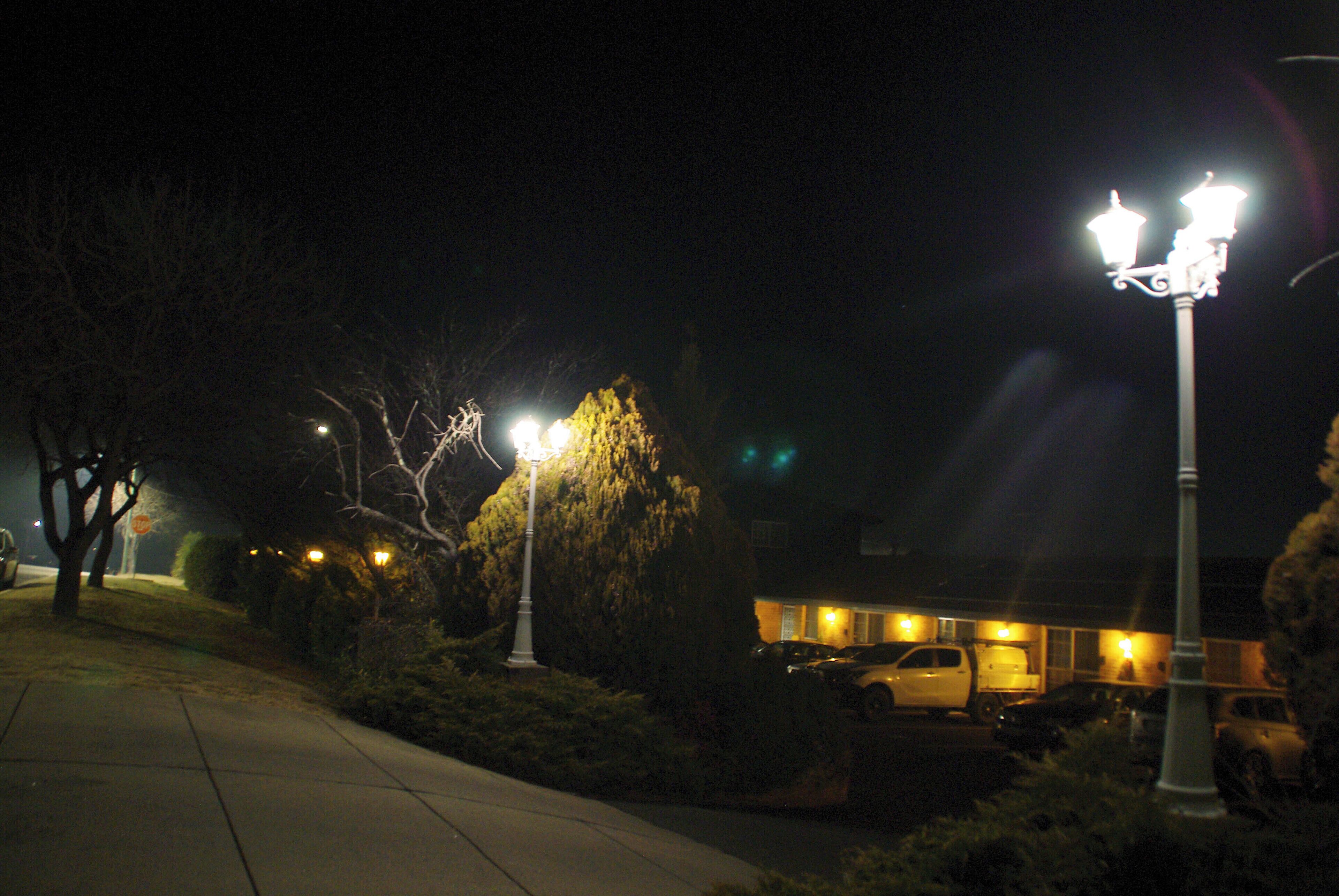 A nighttime street scene with street lamps, parked cars, and a building with illuminated windows.