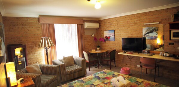 A cozy hotel room with a brick wall, a sofa, a desk with a chair, a television, and a bed with a floral bedspread. - White Lanterns Motel (Armidale)