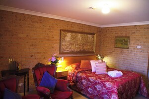 Habitación de hotel con pared de ladrillo, un cuadro grande, cama con edredón estampado con flores, un sillón rojo, una mesita de noche con lámpara y una pequeña mesa con libros y un jarrón de flores.