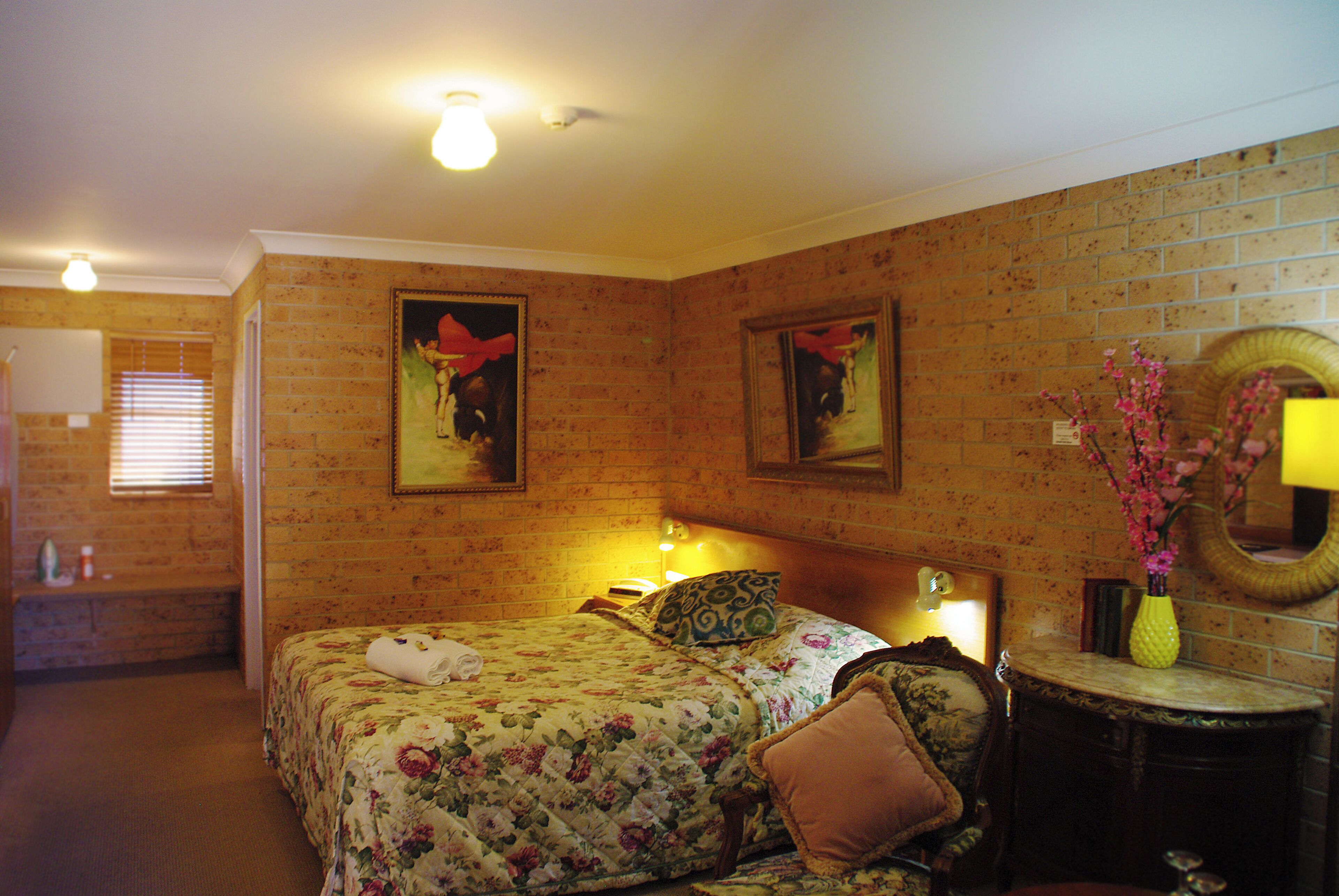 A room with a brick wall, a bed with floral bedding, a nightstand, a mirror, and a vase with flowers.