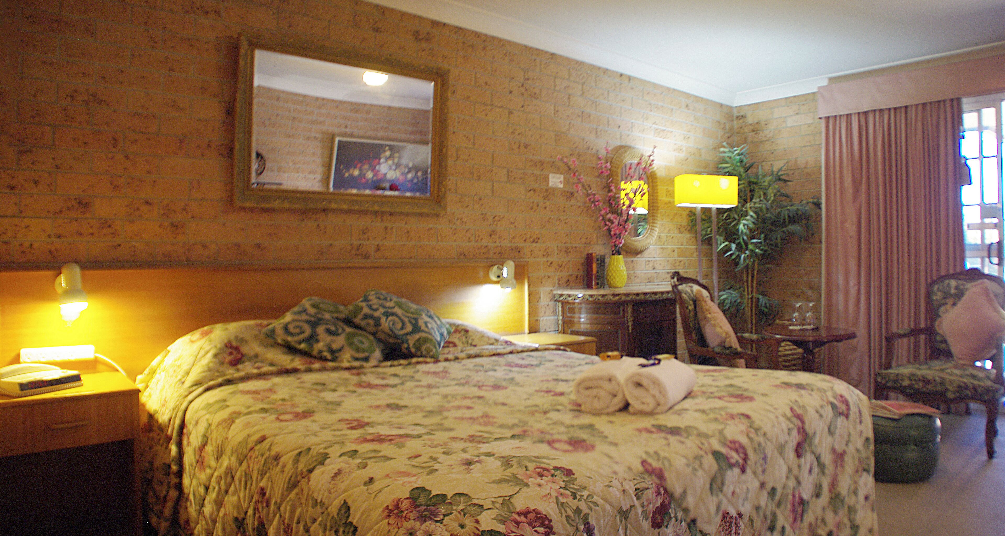 A hotel room with a large bed, a nightstand with a lamp, a chair, a small table, and a mirror on the wall.