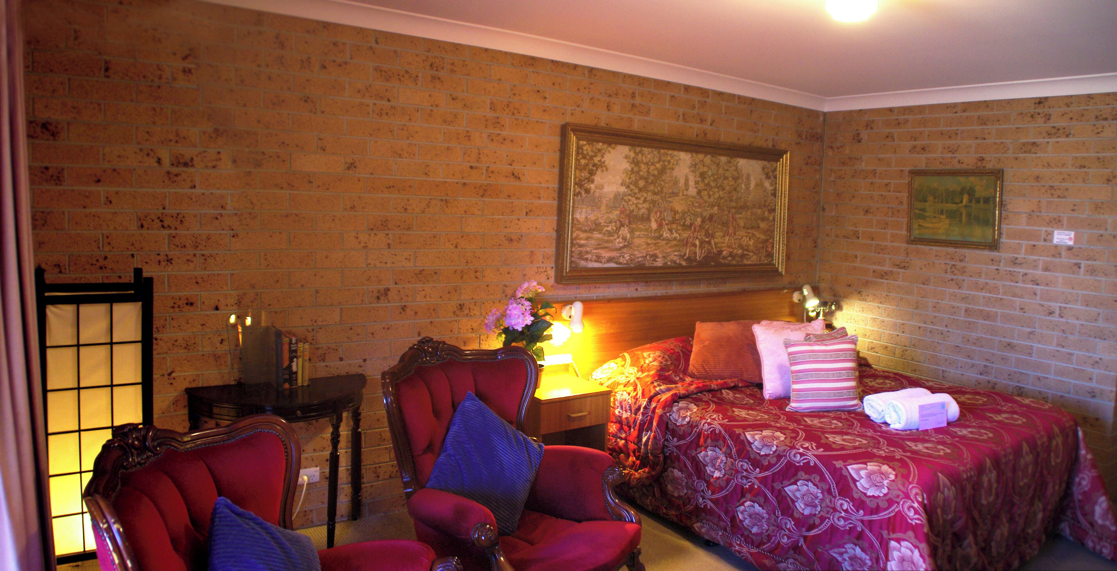 Een kamer met een bakstenen muur, een bed met een bloemenbedekking, een rode fauteuil, een houten nachtkastje en een ingelijst schilderij boven het bed.