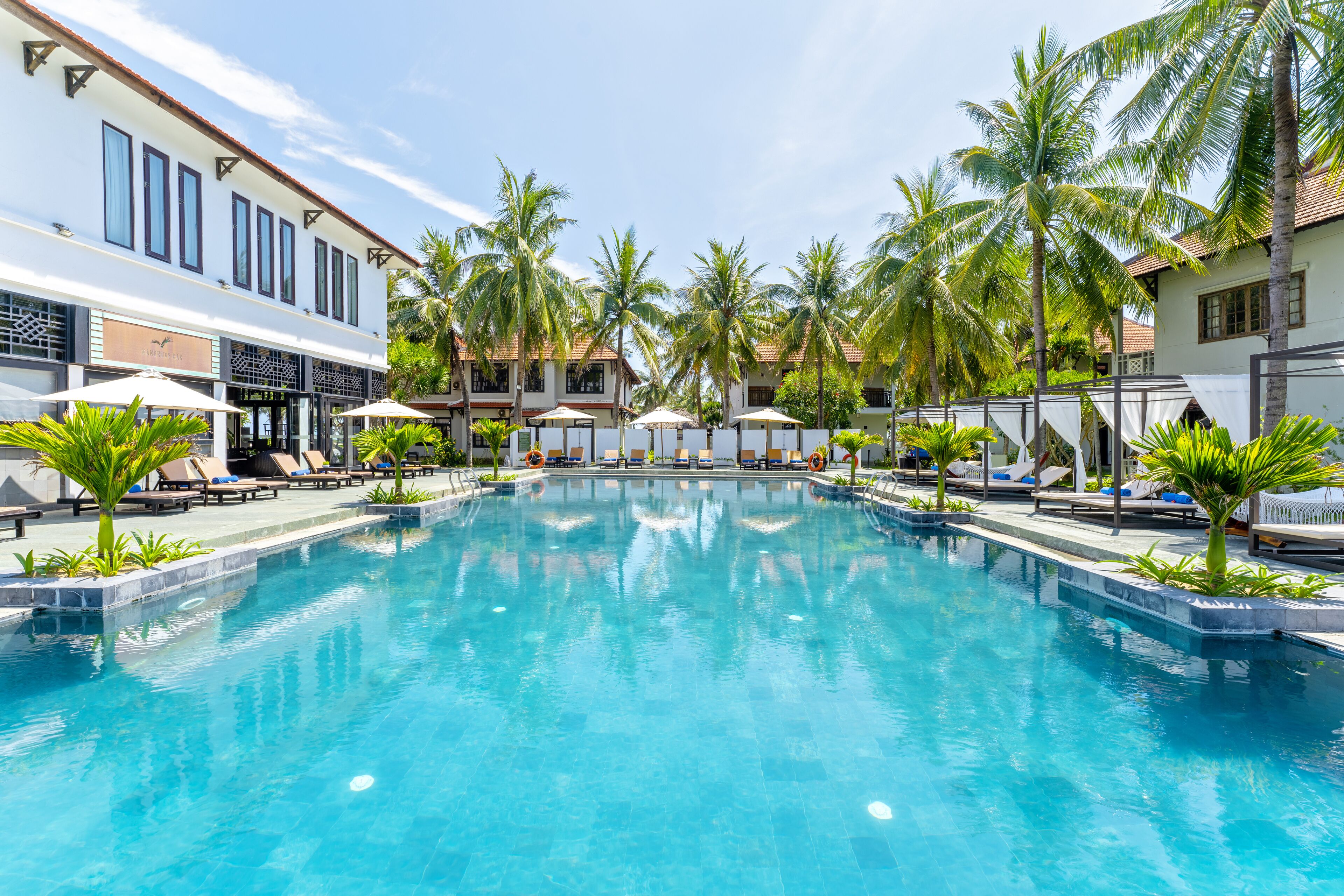 A swimming pool with lounge chairs and umbrellas, surrounded by palm trees and buildings.