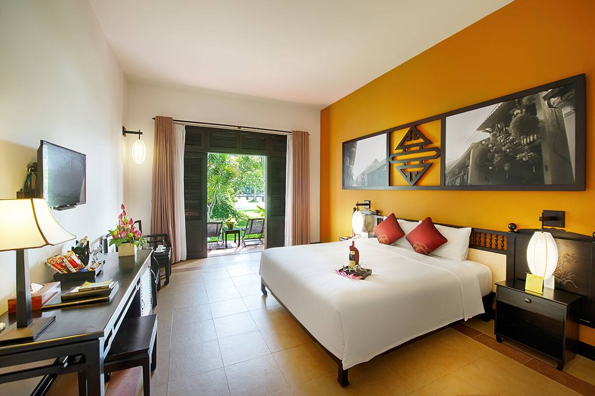 A hotel room with a bed, bedside tables, a desk, a chair, a TV, and a view of the outdoors.