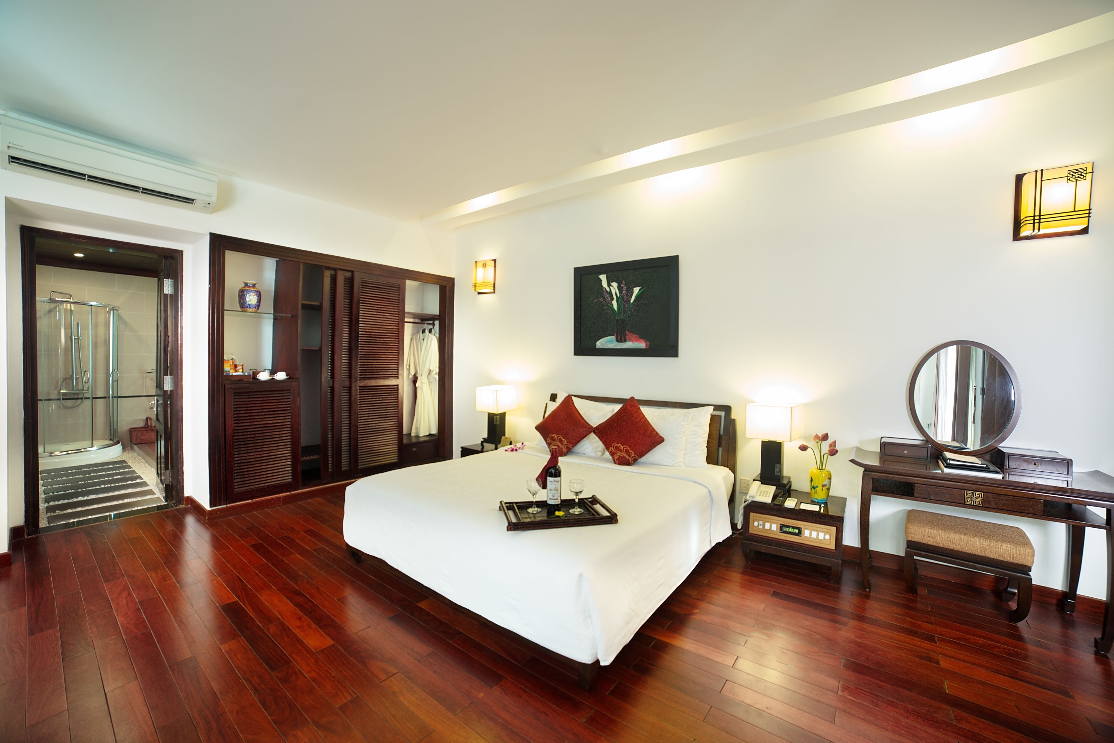 A hotel room with a large bed, a desk with a mirror, a bedside table with a lamp, and a shower area.