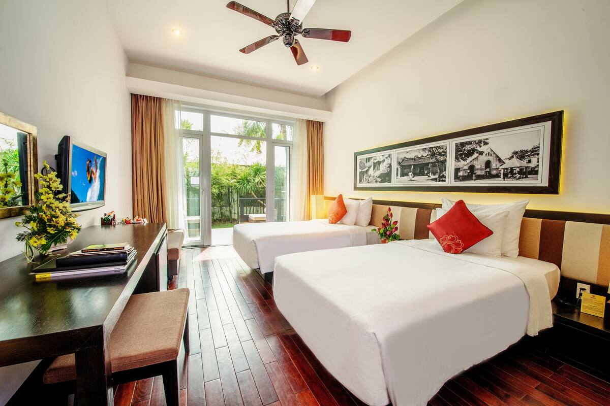 A hotel room with two beds, a desk with a chair, a ceiling fan, and a large window with curtains.