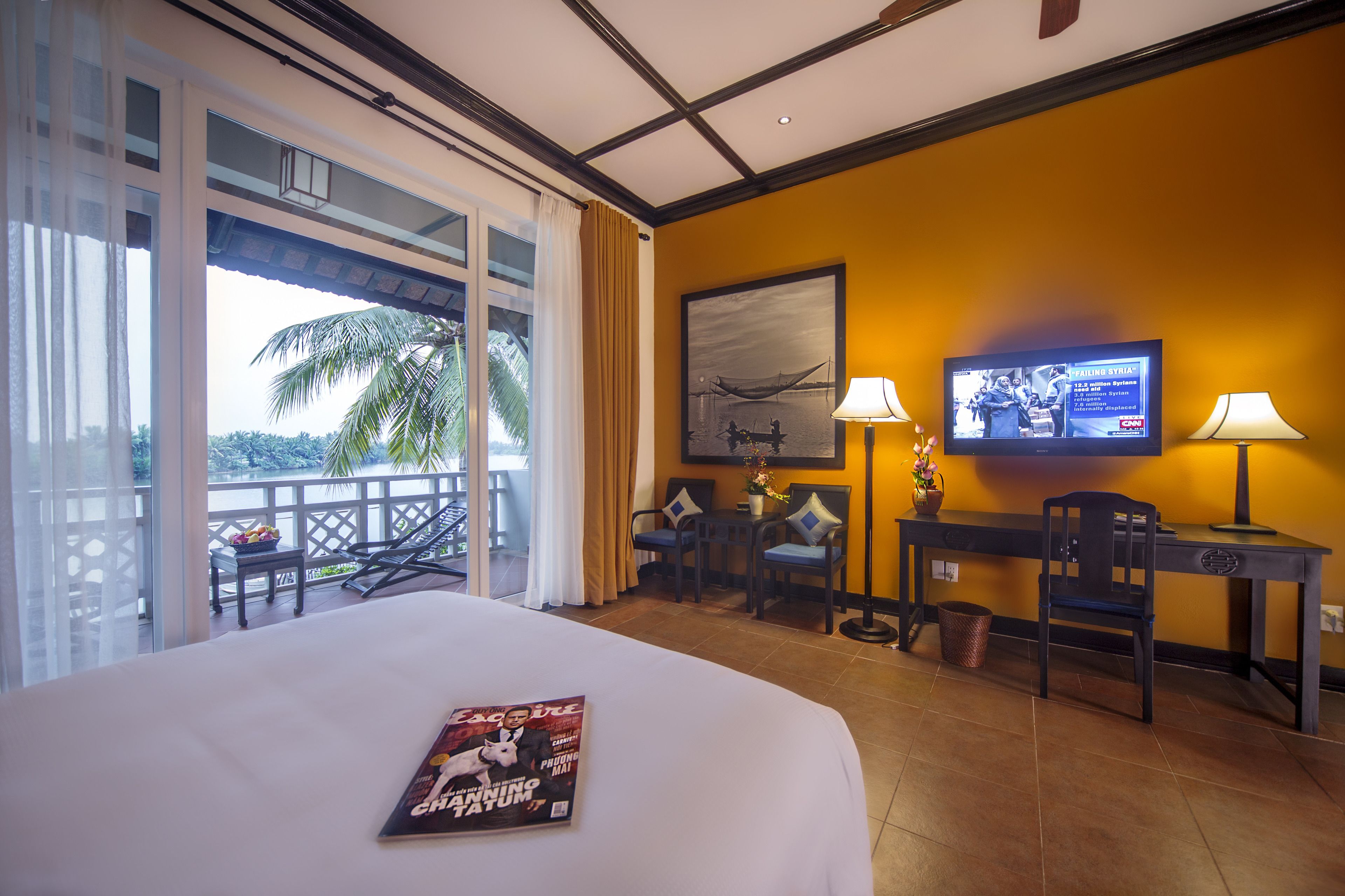 Camera d'albergo con un letto, una scrivania con televisore, un balcone con vista su palme e una rivista sul letto.