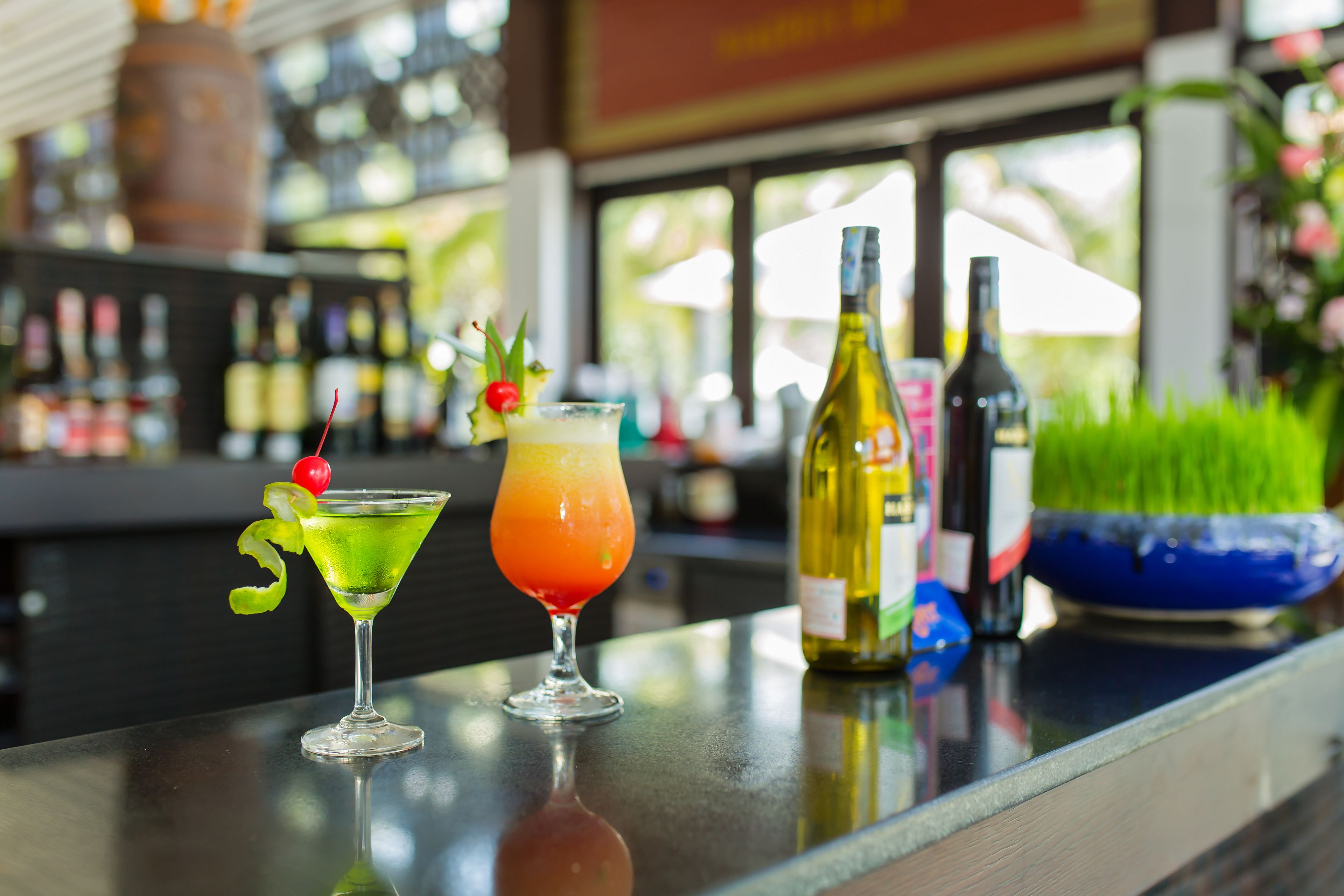 Two cocktails on a bar counter with bottles and a bowl in the background.