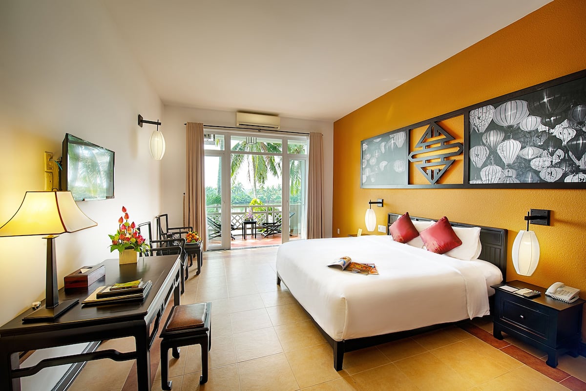 A hotel room with a large bed, a desk with a lamp, a chair, and a view of the outdoors through a glass door.