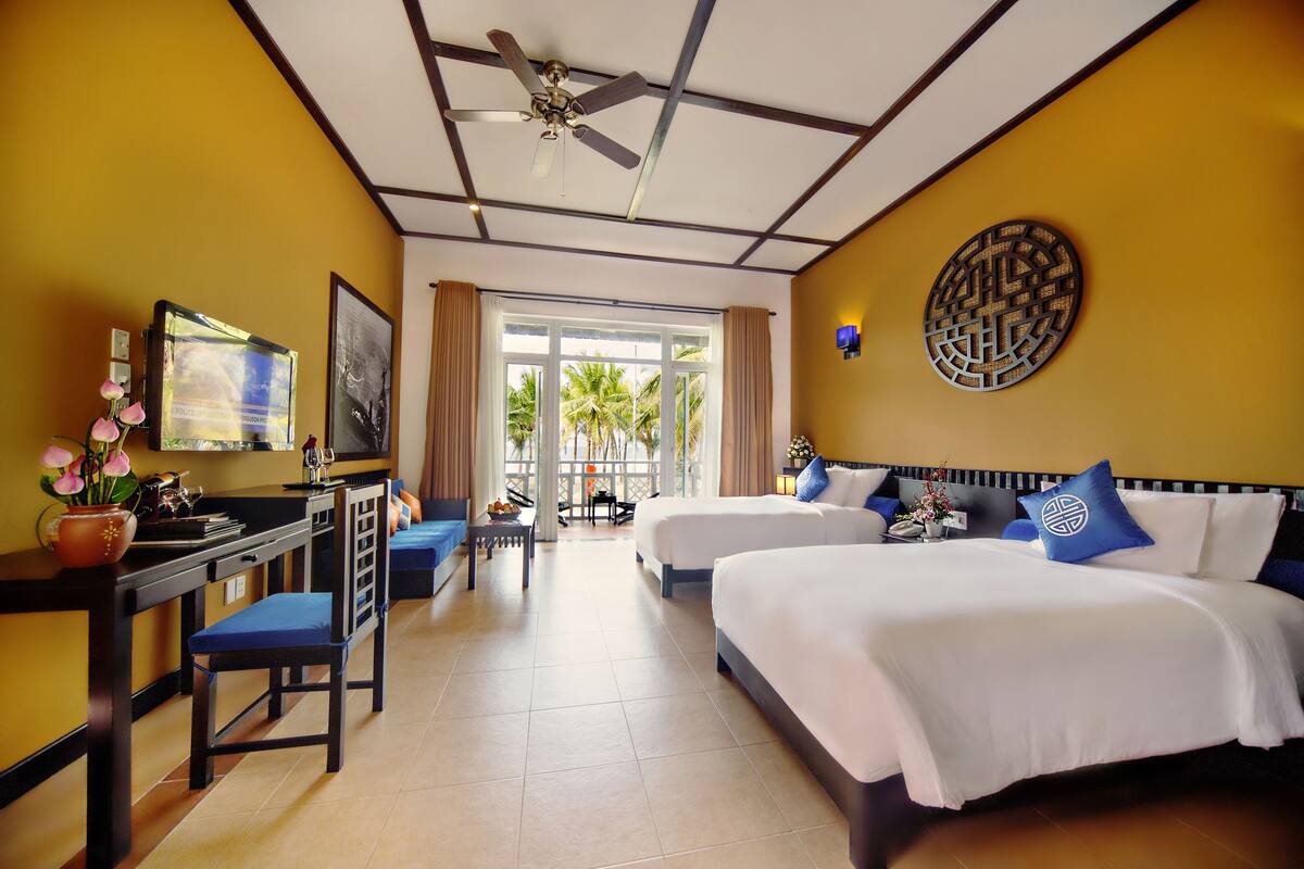 A hotel room with two beds, a desk with a chair, a television, and a large window with curtains.