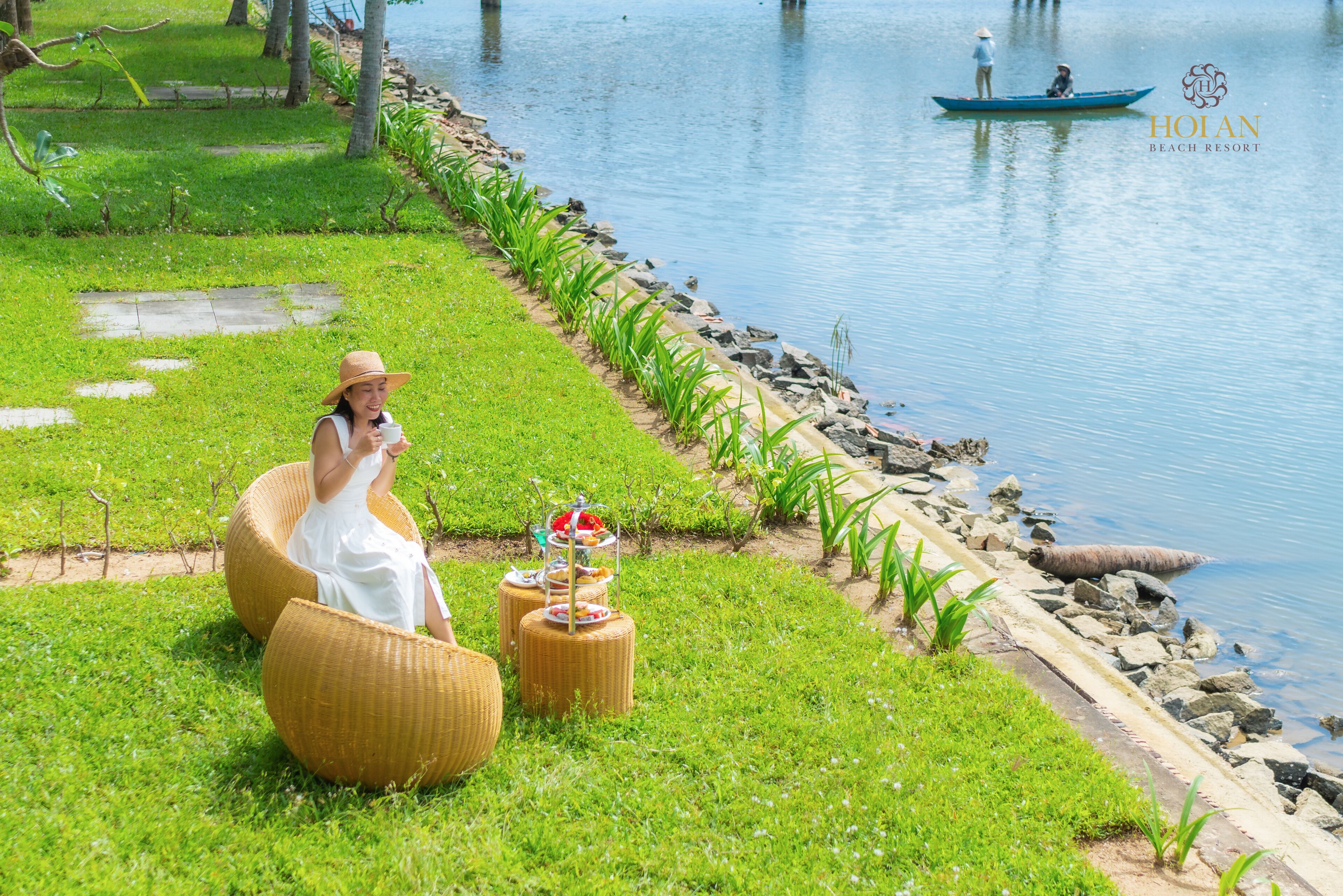 A serene lakeside setting with a woman in a white dress and hat sitting on a wicker chair, a small table with food, and a boat with two people in the background.