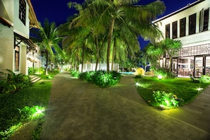A well-lit pathway lined with palm trees and lush greenery, leading to a building with multiple windows and a covered area. - Hoi An Beach Resort (Da Nang)