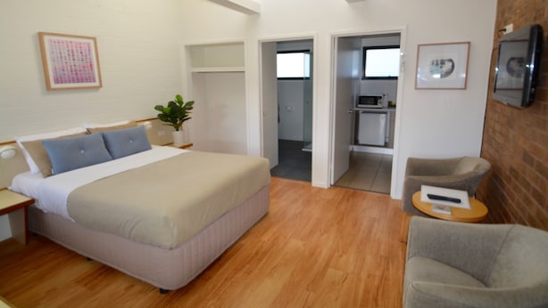 A modern hotel room with a large bed, a seating area with a chair, and a kitchenette in the background.
