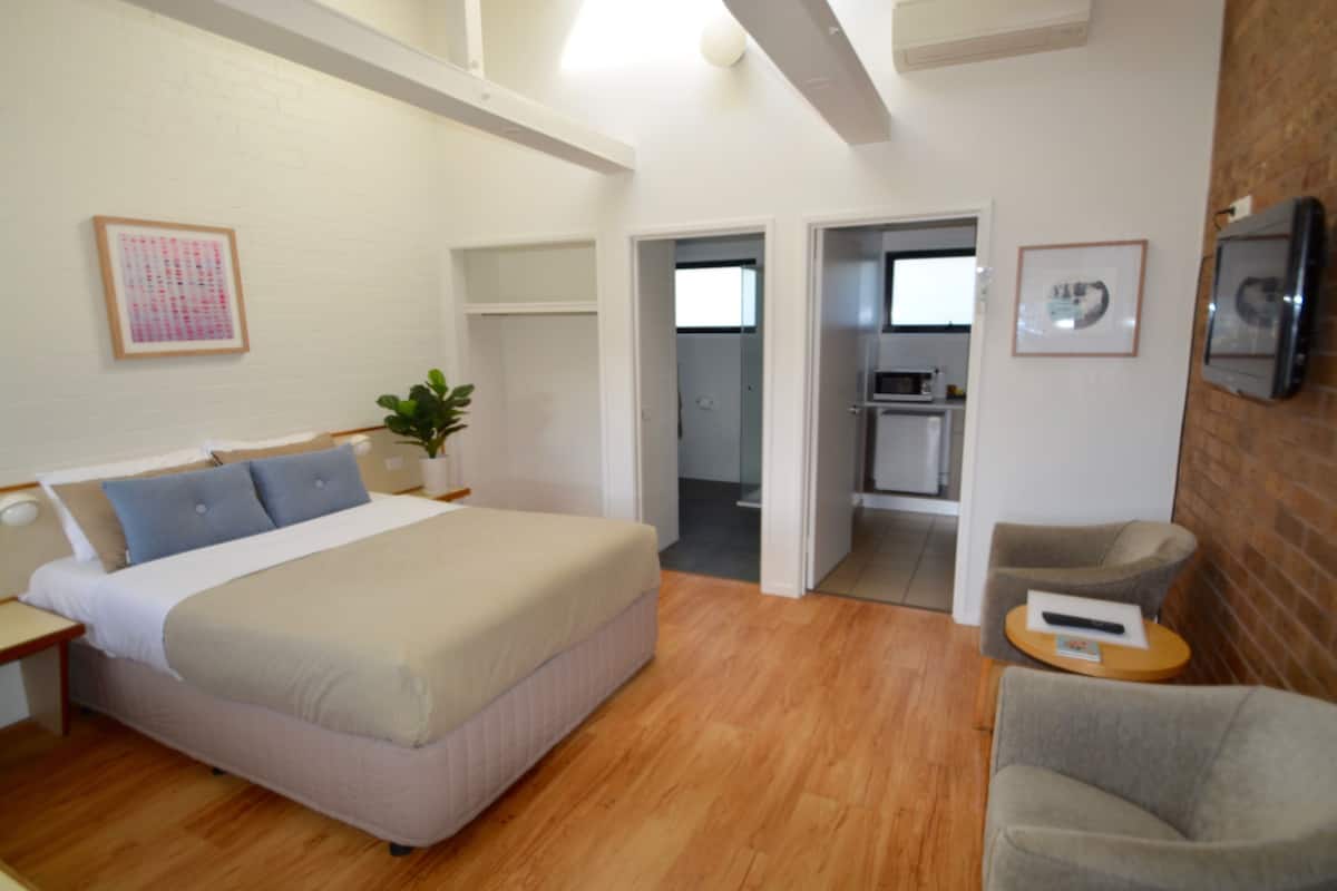 A modern hotel room with a large bed, a seating area with a chair, and a kitchenette in the background.