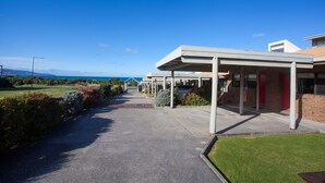 A covered walkway leading to a building with a modern architectural style, a clear sky, and a view of the sea in the background.