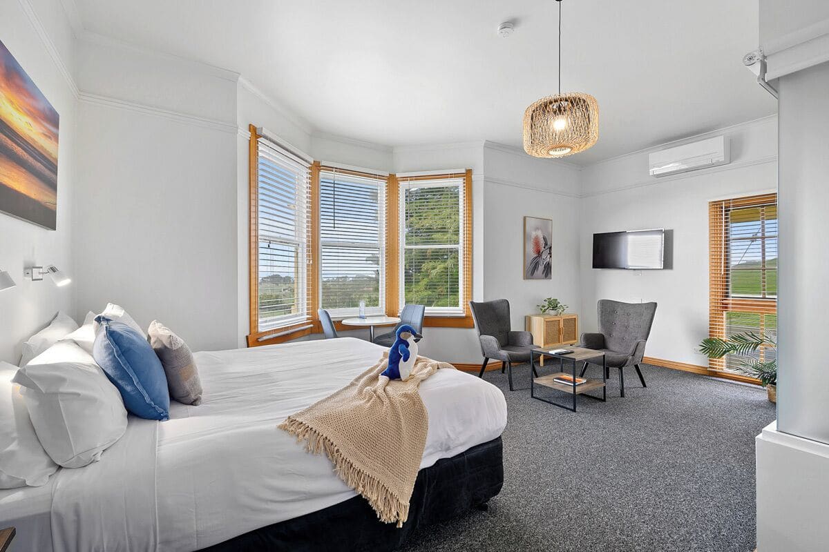 A modern hotel room with a large bed, a desk, two chairs, a television, and large windows with blinds.