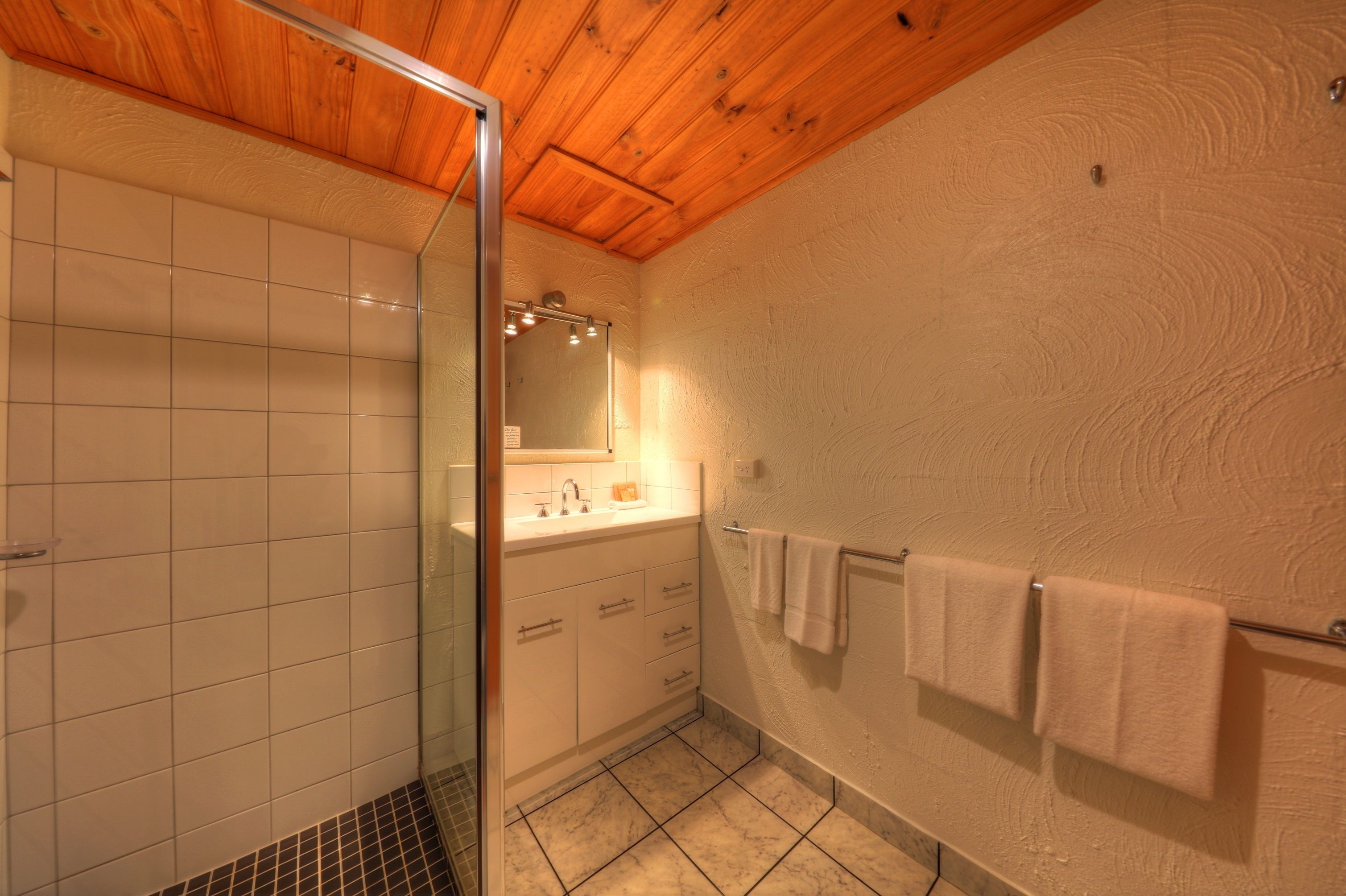 Un baño con cabina de ducha de vidrio, azulejos blancos, techo de madera y un perchero con toallas.