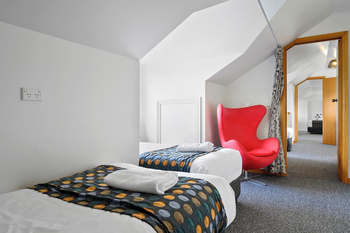 Una habitación de hotel con dos camas, una silla roja y vista hacia otra habitación.