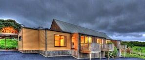 A wooden cabin with a balcony and a covered porch, surrounded by a gravel driveway and a grassy landscape.