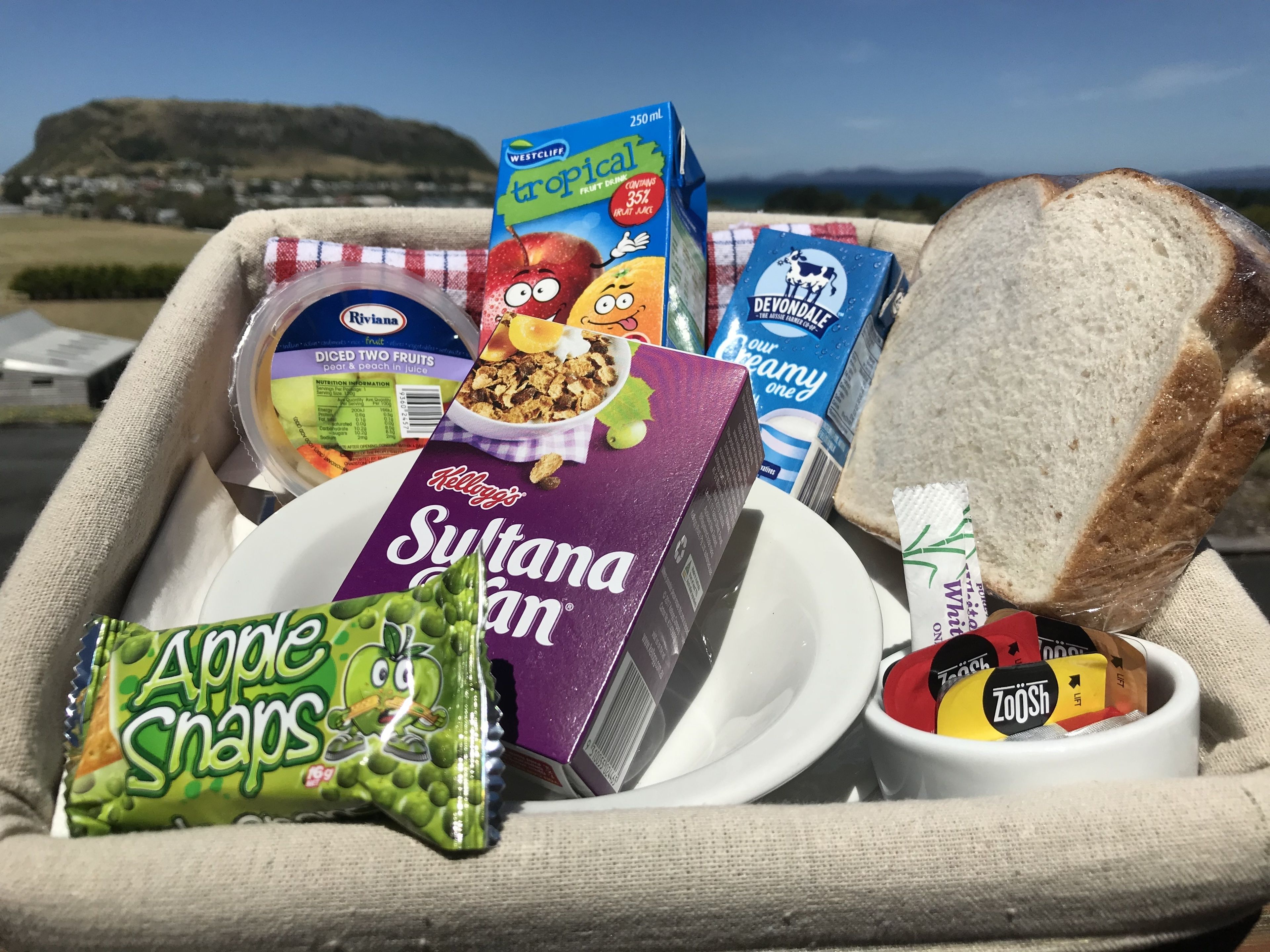 A picnic basket with various food items including a box of Kellogg's Sultana Cereal, a pack of apple snaps, a container of yogurt, a box of Westcliff Tropical Fruit Juice, a loaf of bread, and a tube of chocolate spread.