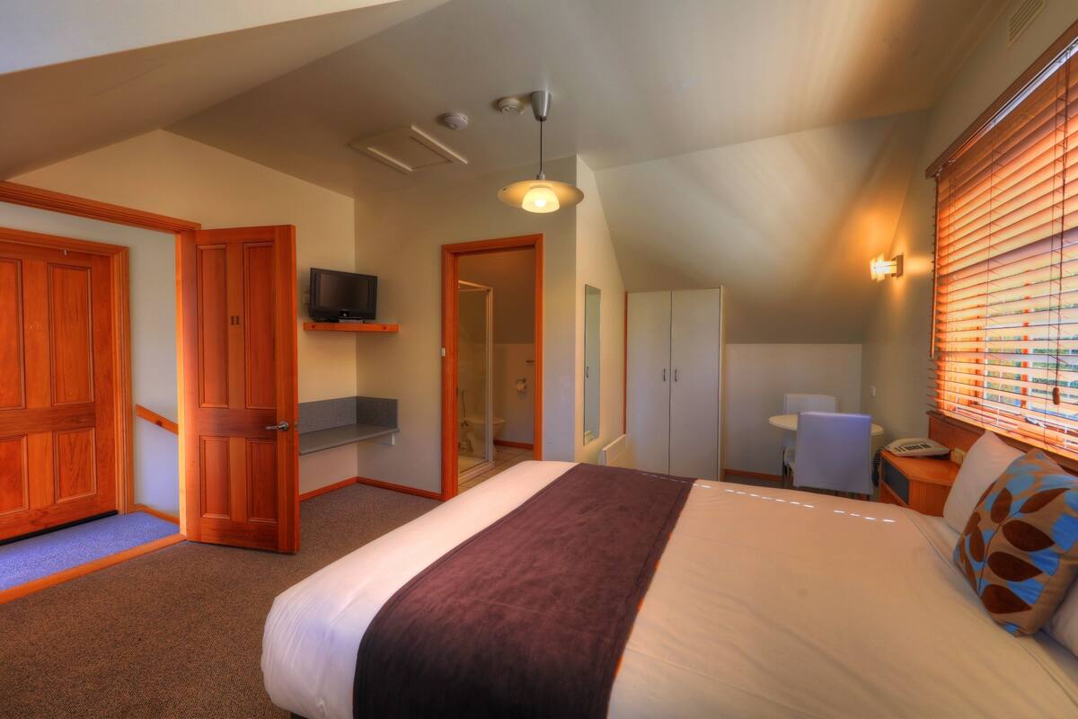 A bedroom with a bed, wooden wardrobe, a television, a desk, and a window with blinds.