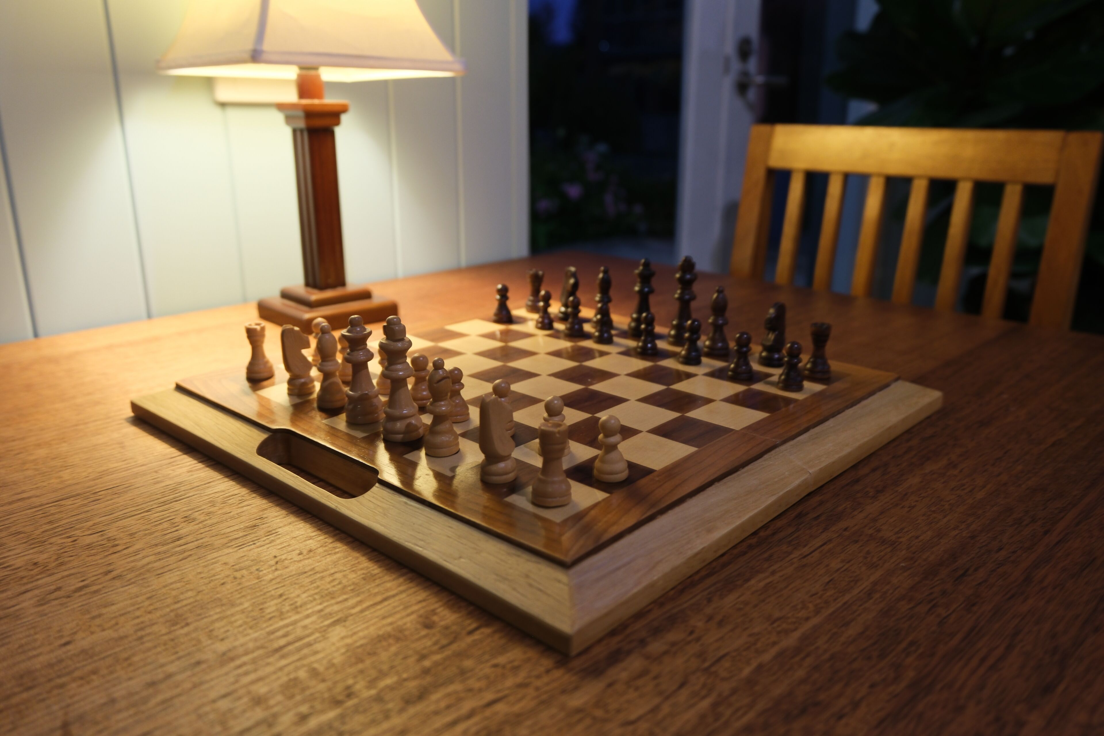 A wooden chessboard on a table with a lamp in the background.