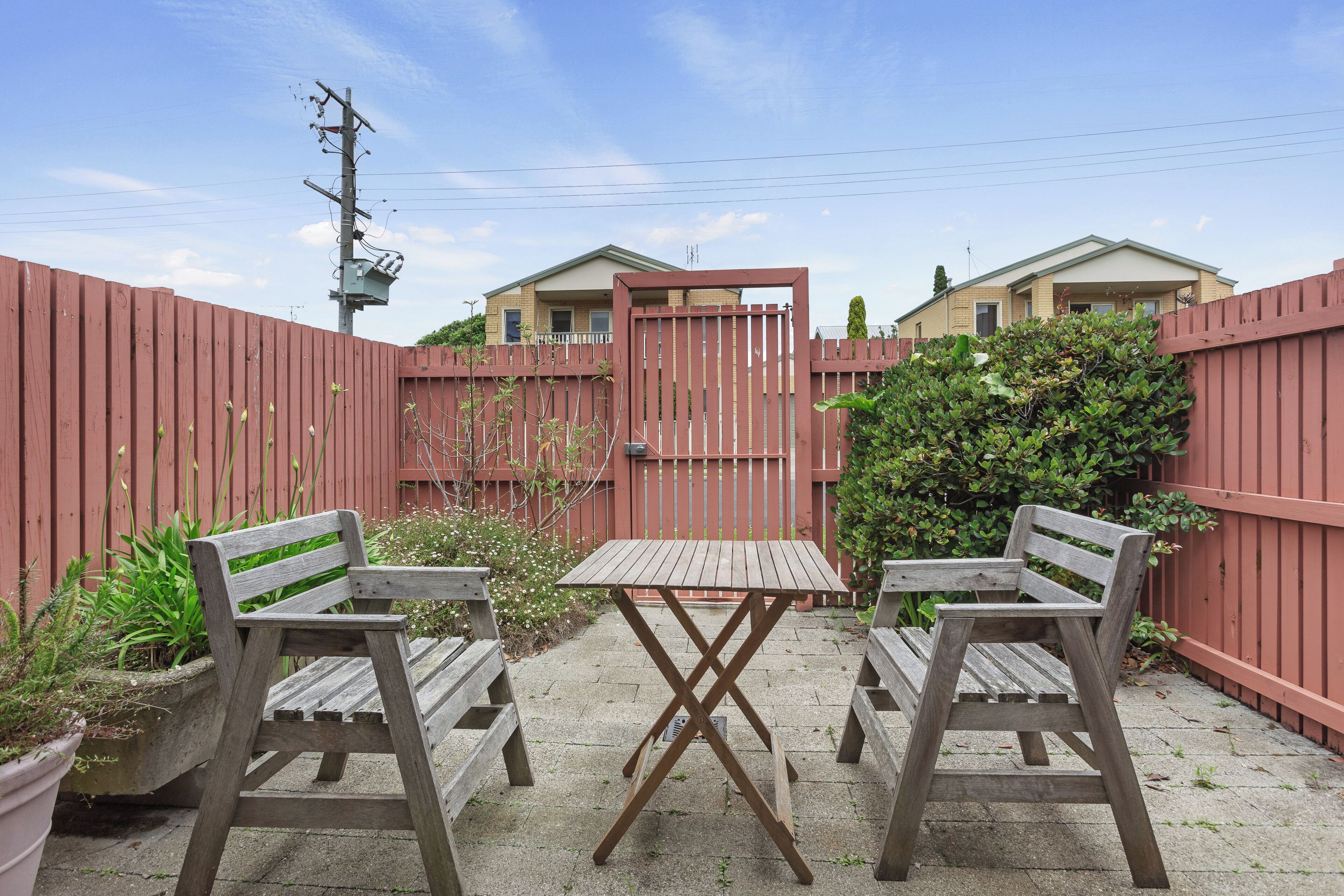 Une terrasse avec deux chaises en bois et une table, entourée d’une clôture rouge, avec des maisons en arrière-plan.