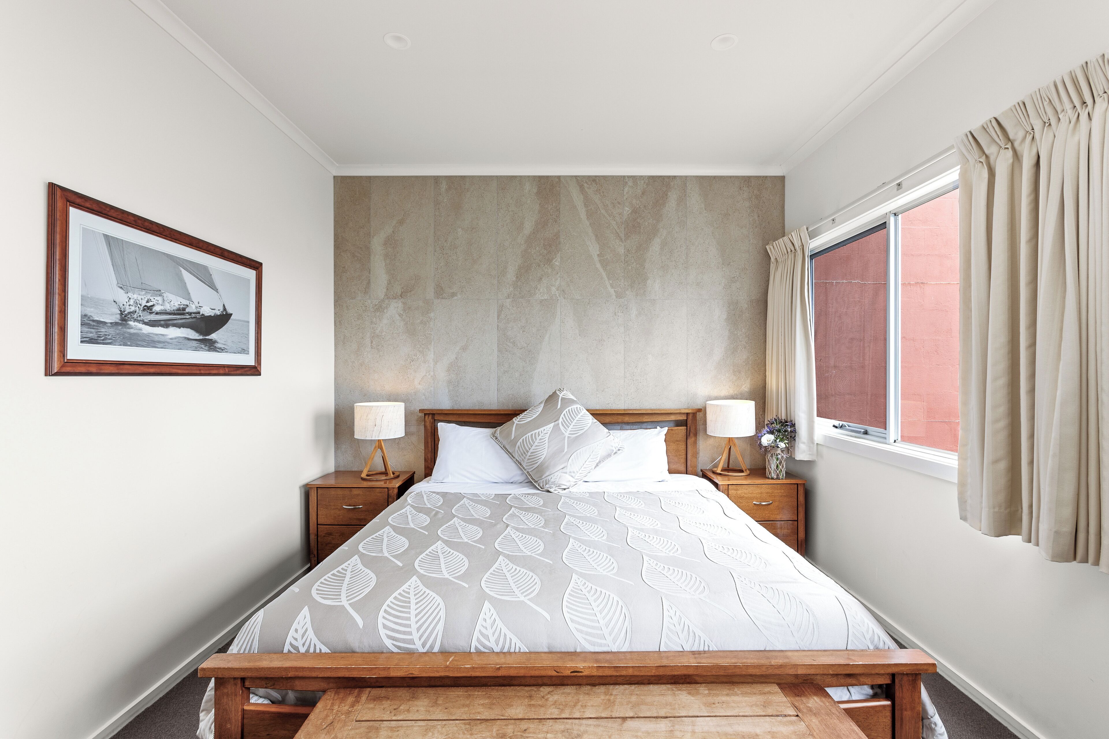 Ein Schlafzimmer mit Bett, Holz-Kopfteil, Nachttischen mit Lampen, einem gerahmten Bild an der Wand und einem Fenster mit Vorhängen.