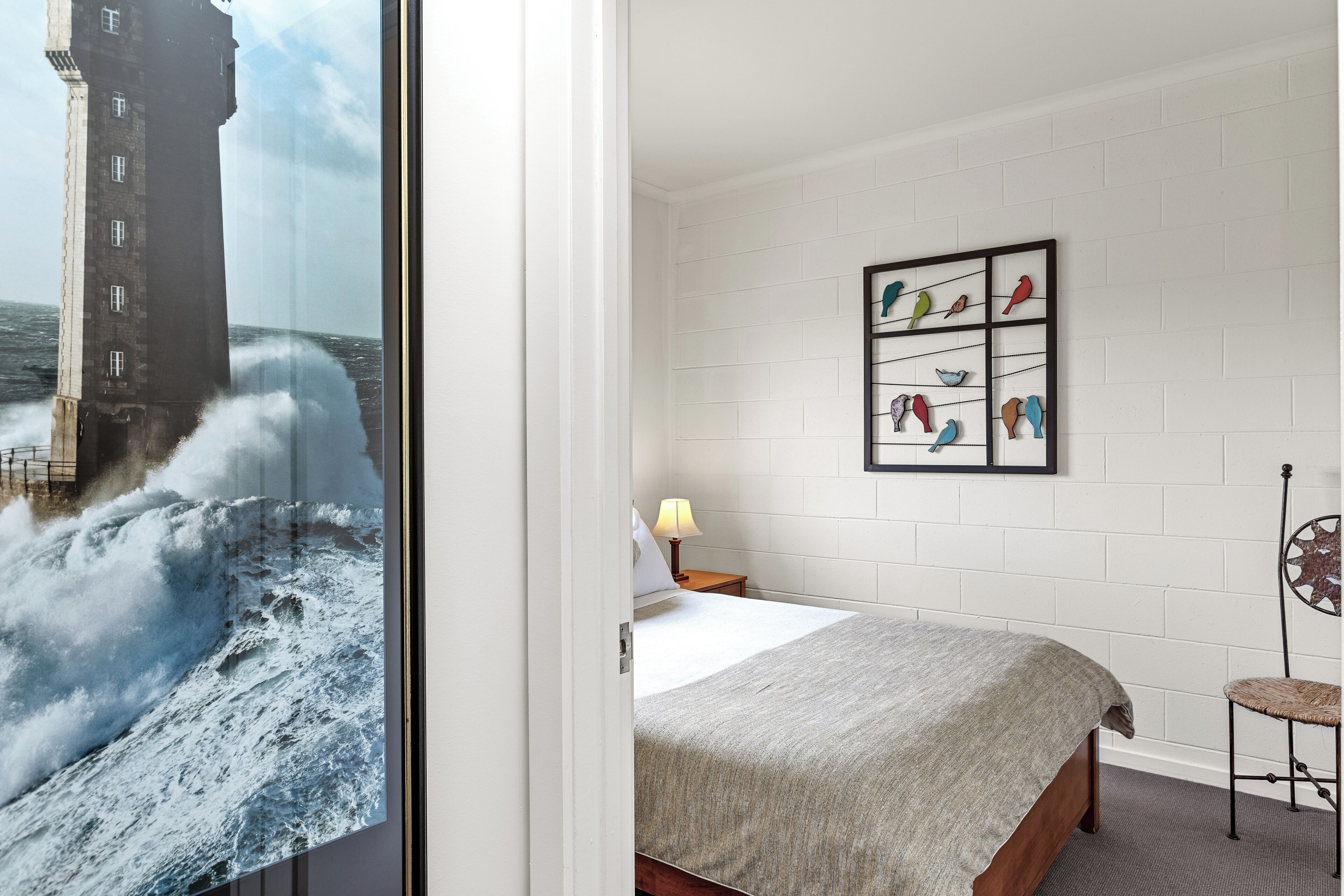 Et hotelværelse med en seng, et natbord med en lampe, en stol og en væg med et billede af fugle i ramme.
