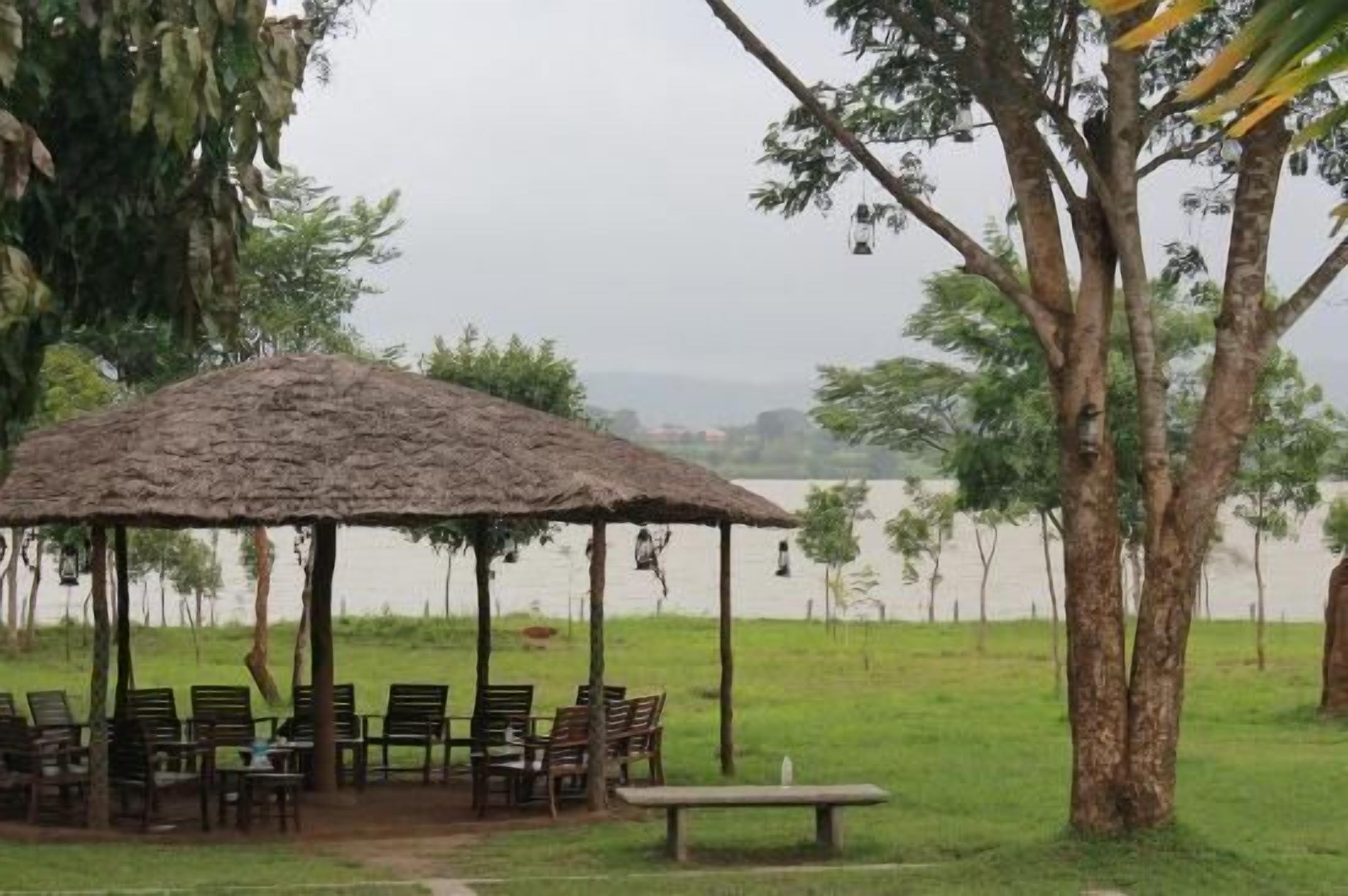 A thatched-roof pavilion with wooden chairs and a bench, surrounded by trees and a body of water.