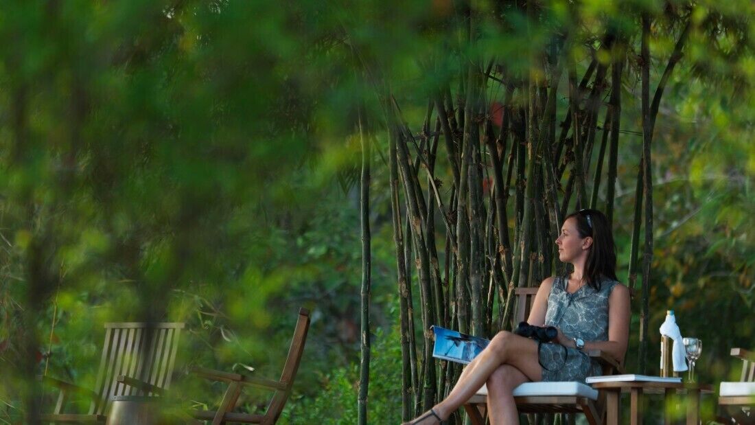 A woman seated on a wooden chair outdoors, holding a camera and a notebook.
