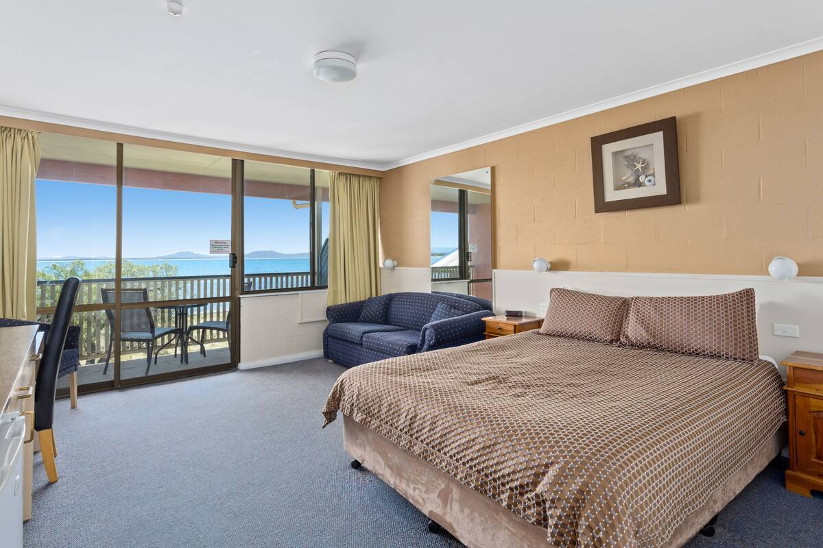 A hotel room with a large bed, a desk, and a balcony with a view of the sea.