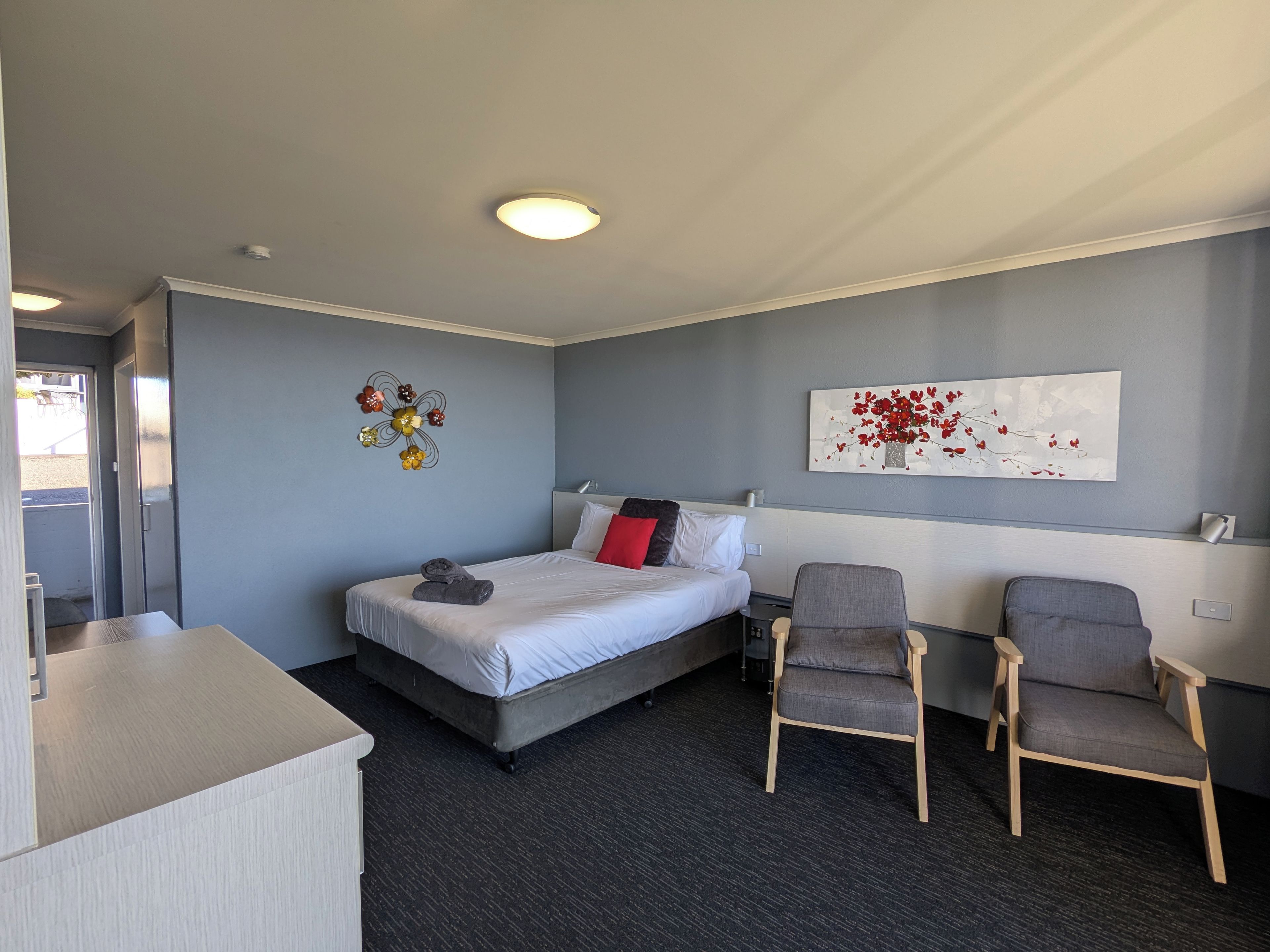 Quarto de hotel com cama, duas cadeiras, escrivaninha, uma obra de arte na parede e luminária de teto.