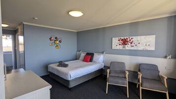 Quarto de hotel com cama, duas cadeiras, escrivaninha, uma obra de arte na parede e luminária de teto.