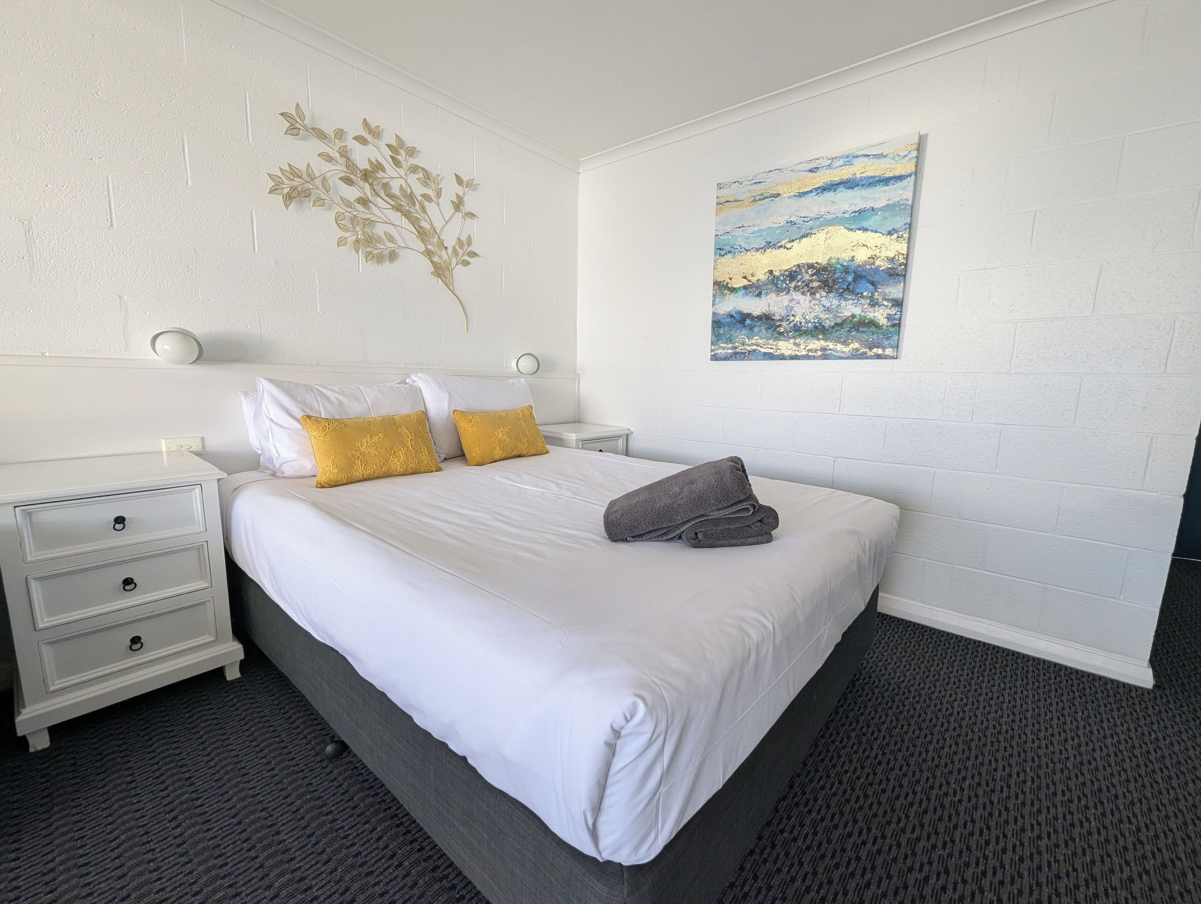 Una habitación de hotel con una cama, mesitas de noche blancas, una lámpara de pared y un cuadro.