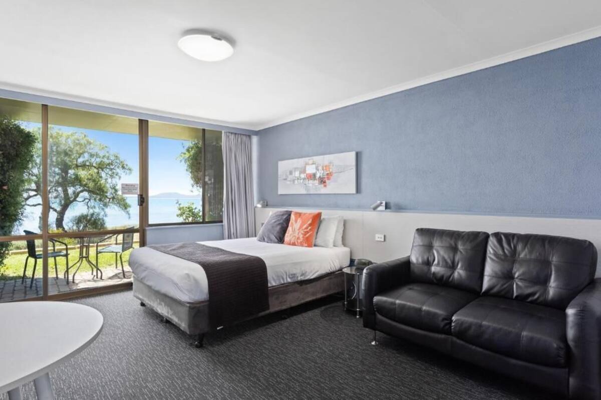A modern hotel room with a bed, a black leather sofa, a small table, and a balcony with a view of trees and a body of water.