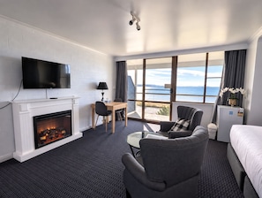 A hotel room with a fireplace, a TV, a desk, a chair, a bed, and a view of the sea. - Swansea Motor Inn (Swansea)